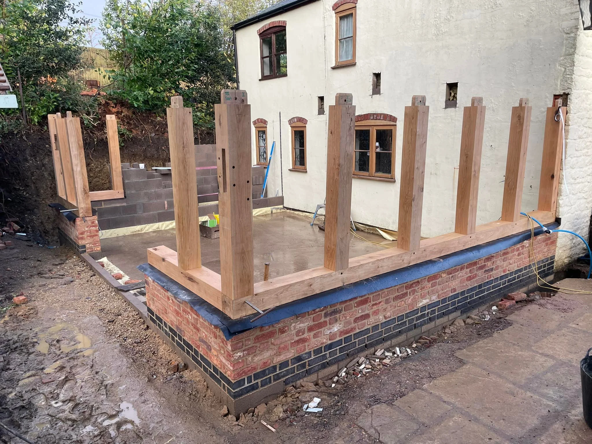 Construction site with wooden posts and brick foundation, possibly for a building or deck, in front of a residential house.