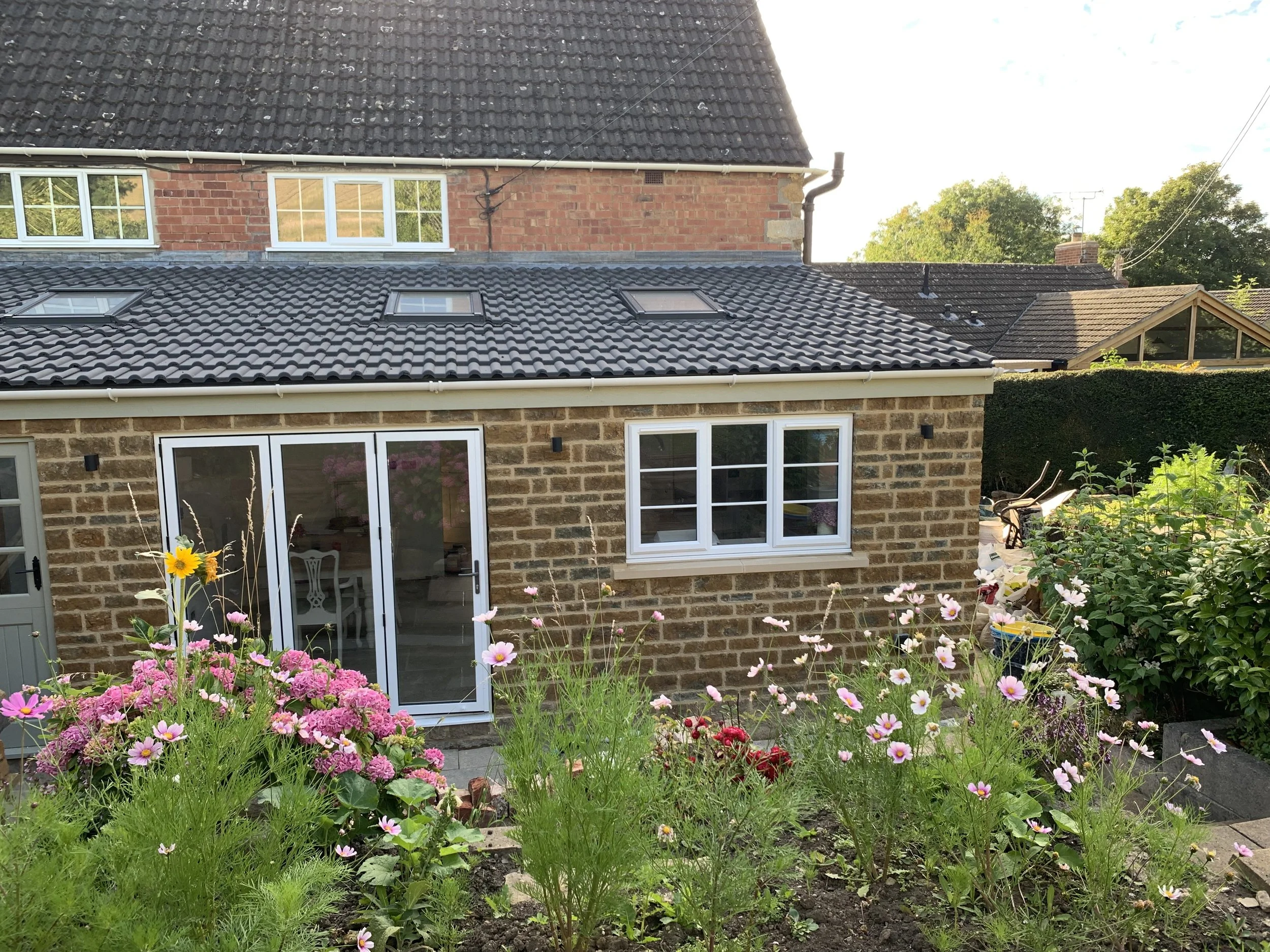 View of a backyard with flowering plants in front of a brick house that has glass double doors and a window. The house has a tiled roof with skylights and neighboring houses are visible in the background.