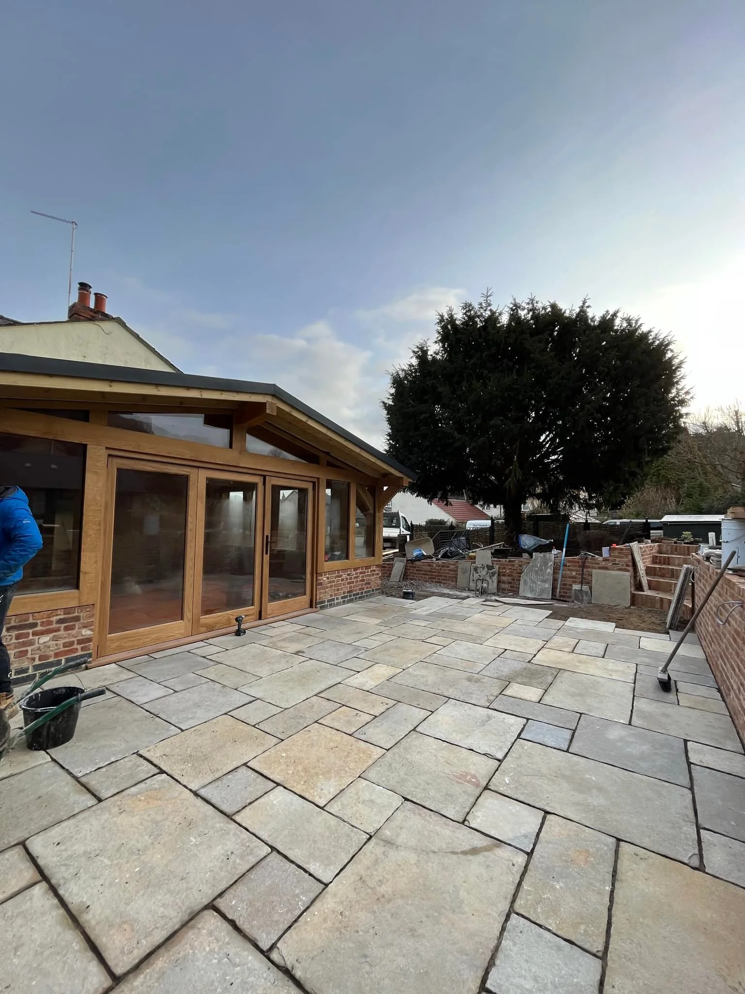 A backyard patio with newly laid stone paving, a wooden extension to a house with glass sliding doors, and a large tree in the background.