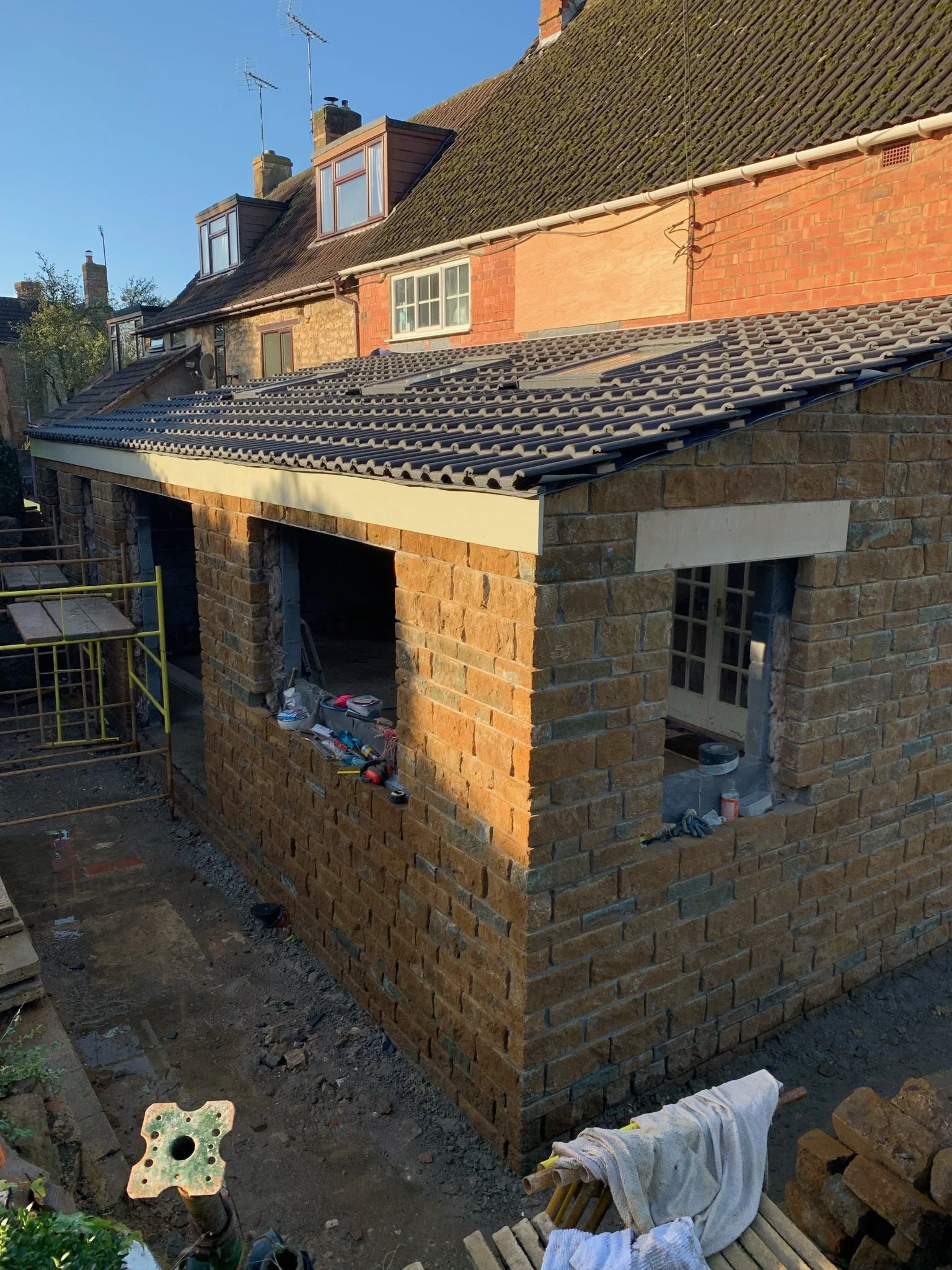 Construction of a brick extension with a tiled roof on a residential building, showing brick walls, windows, and construction tools.