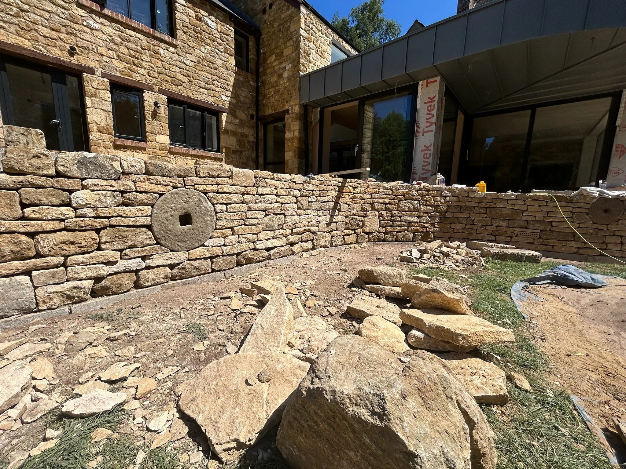 Under construction backyard with stone wall and patio extension, construction materials scattered on ground, partially built structure with windows, and forested area in background.