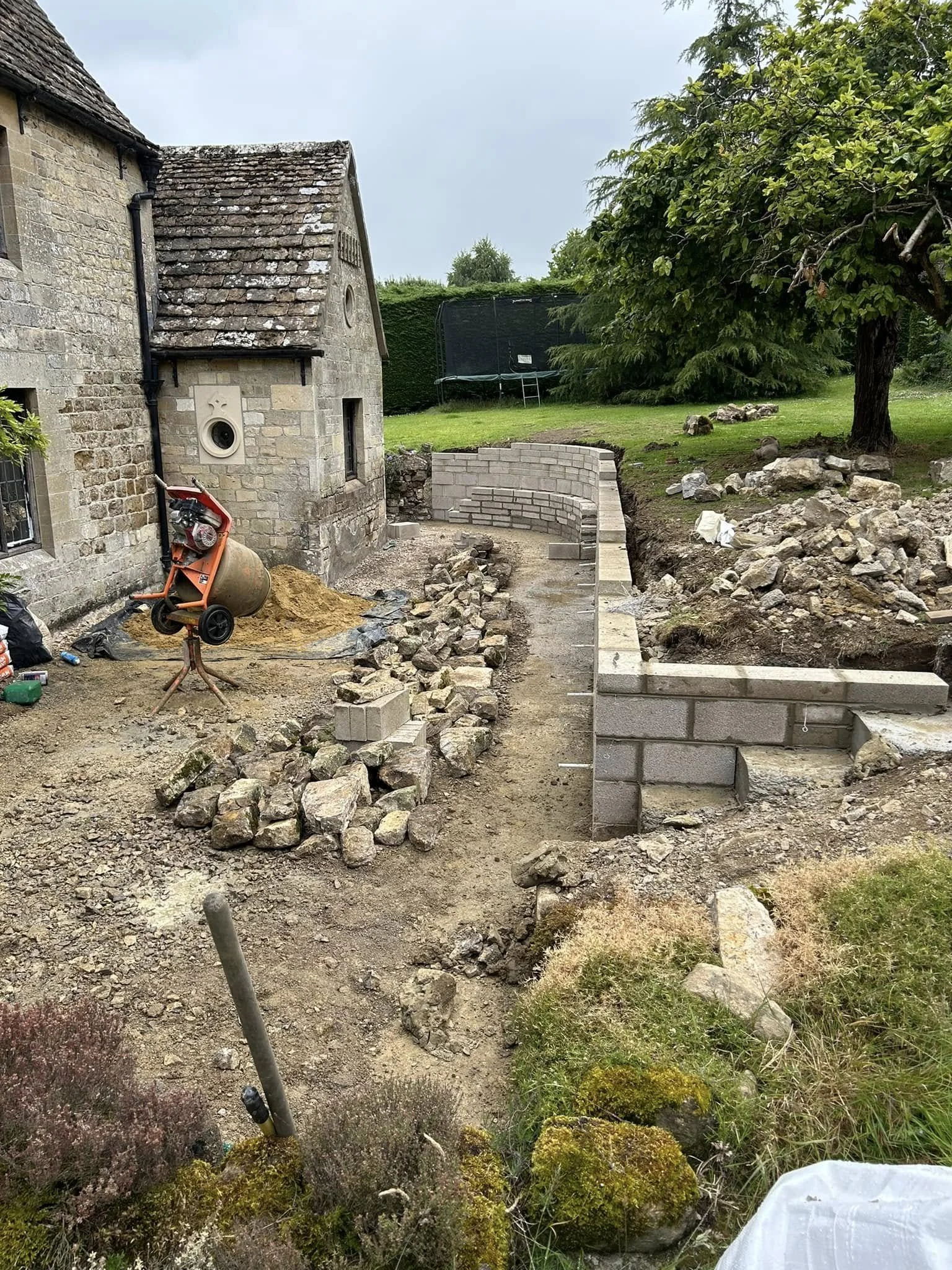 A garden area with construction work in progress, showing a stone wall being built, a cement mixer, and various construction materials, with trees and a lawn in the background.
