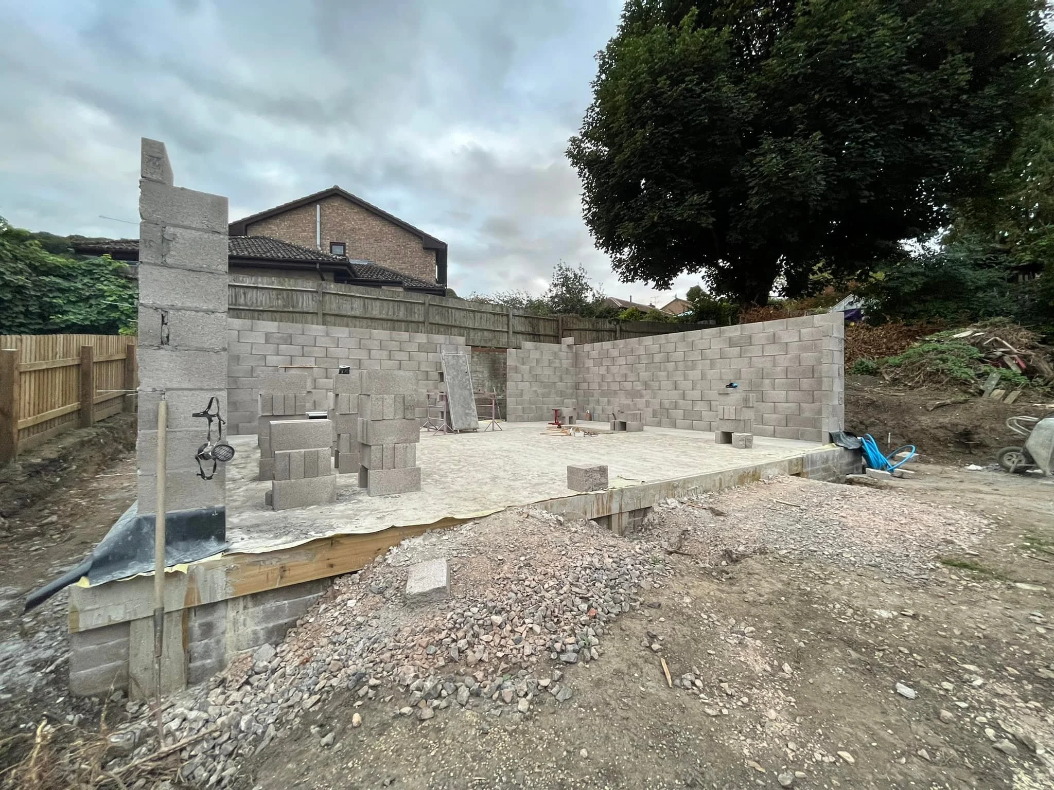 Construction site with partially built cinder block walls, a concrete floor, and construction materials including stacks of cinder blocks and tools around the site, with a wooden fence and trees in the background under a cloudy sky.