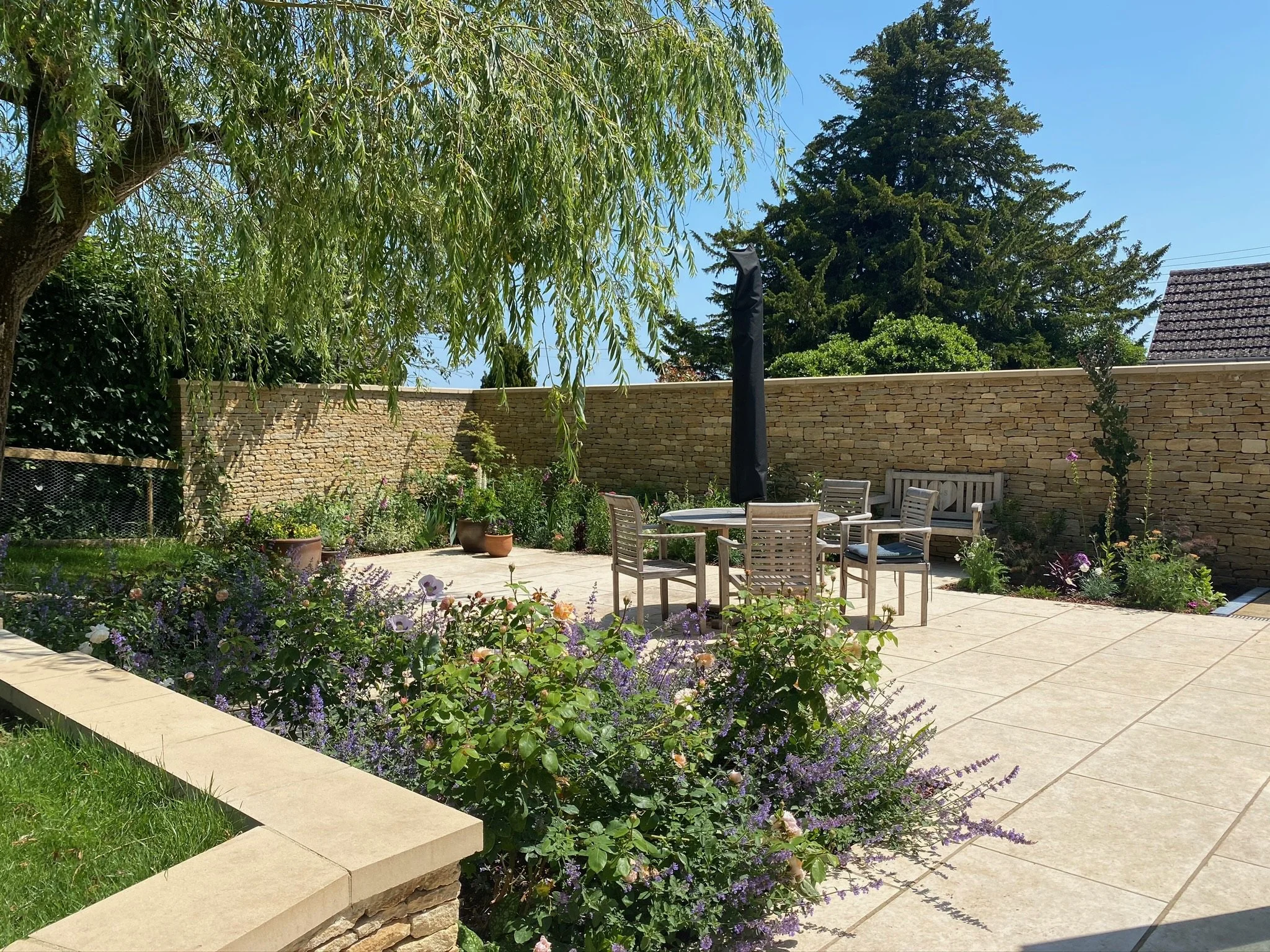 A sunny backyard patio with outdoor furniture, potted plants, a stone wall, and lush green trees.