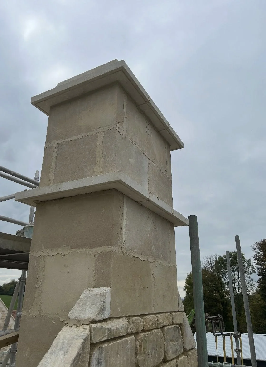 Close-up of a stone chimney under construction or renovation, with scaffolding and trees in the background.
