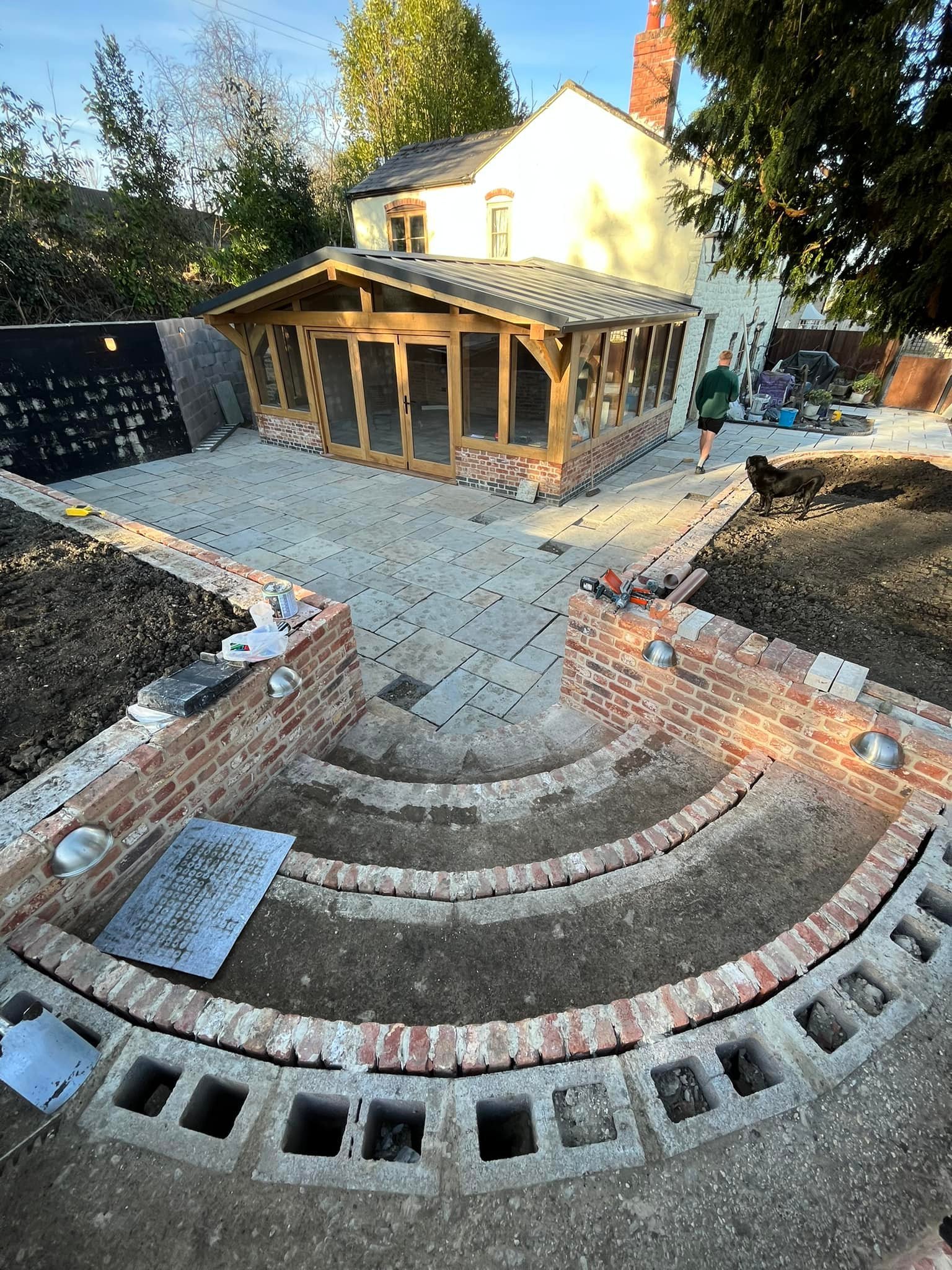 Backyard patio under construction with stone pavers, a brick and cinder block wall, and a sunroom attached to a two-story house. A person walks away with a dog nearby.