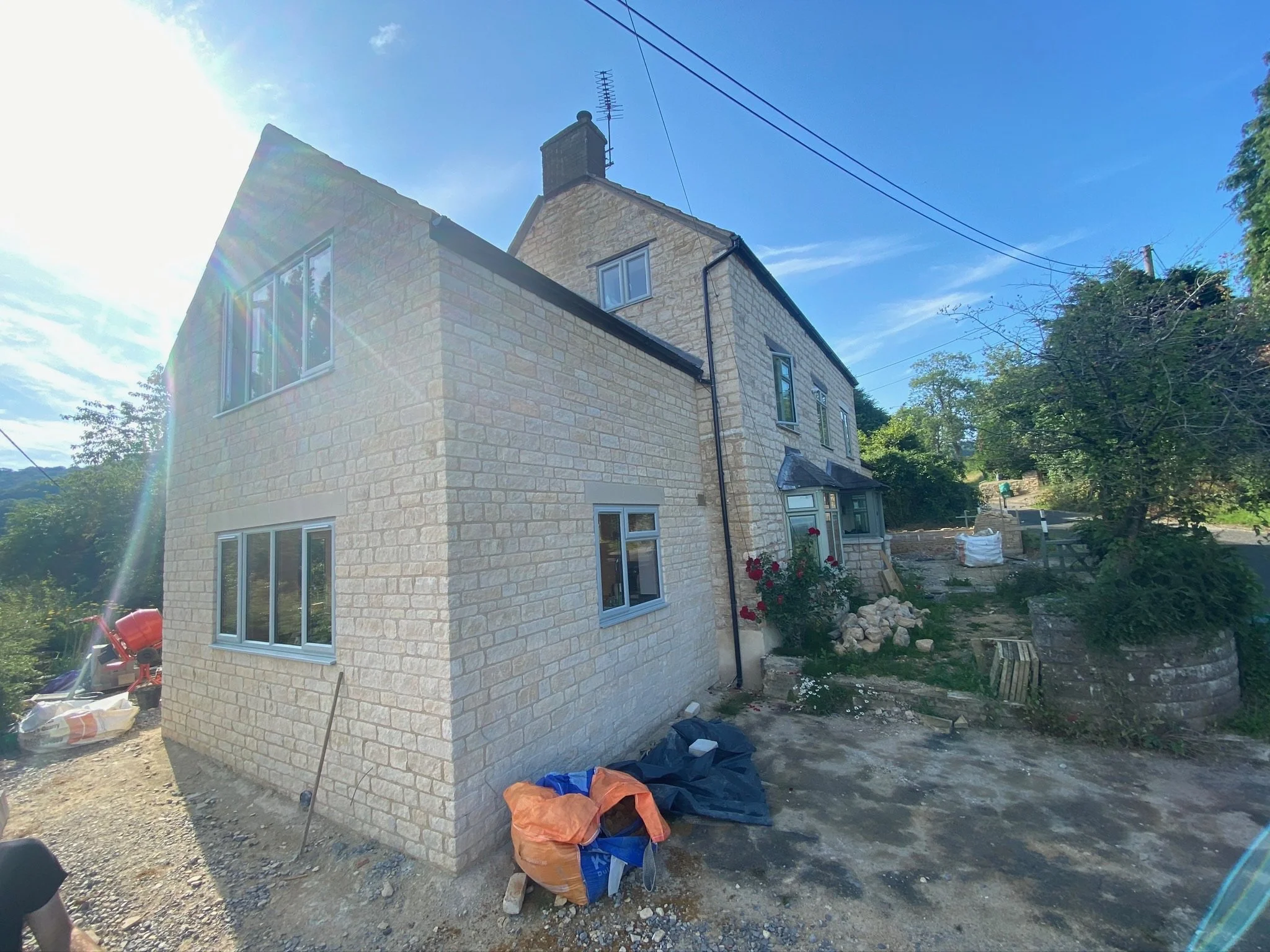 A two-story house under construction with beige brick exterior, multiple white-framed windows, and a steep gable roof, set against a bright blue sky with sunlight glare.