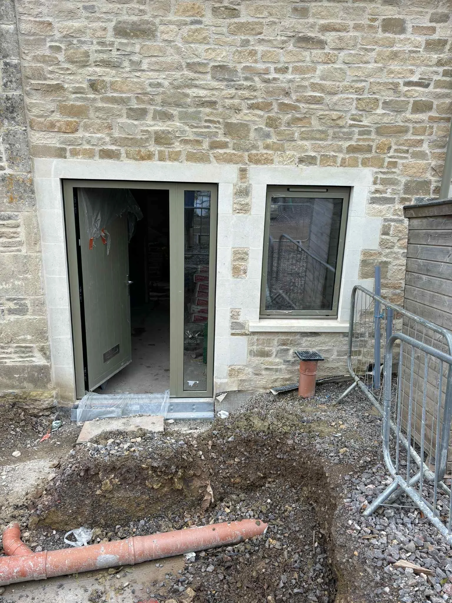 Construction site showing excavated soil near the back door of a brick building, with a partially completed pathway, a pipe protruding from the ground, and metal barriers surrounding the area.