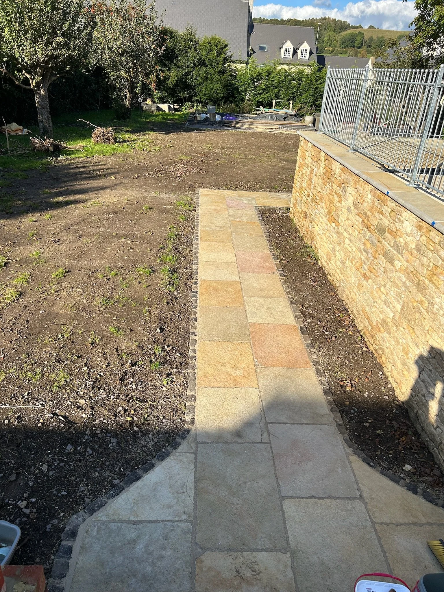 Sidewalk leading away from a stone wall with a metal fence on top, with a garden area and trees in the background on a sunny day.