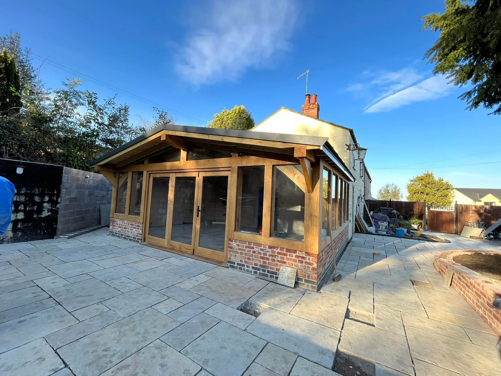 Backyard patio with newly built enclosed wooden sunroom, paved stone surface, and a white house with brick chimney in the background on a clear day.