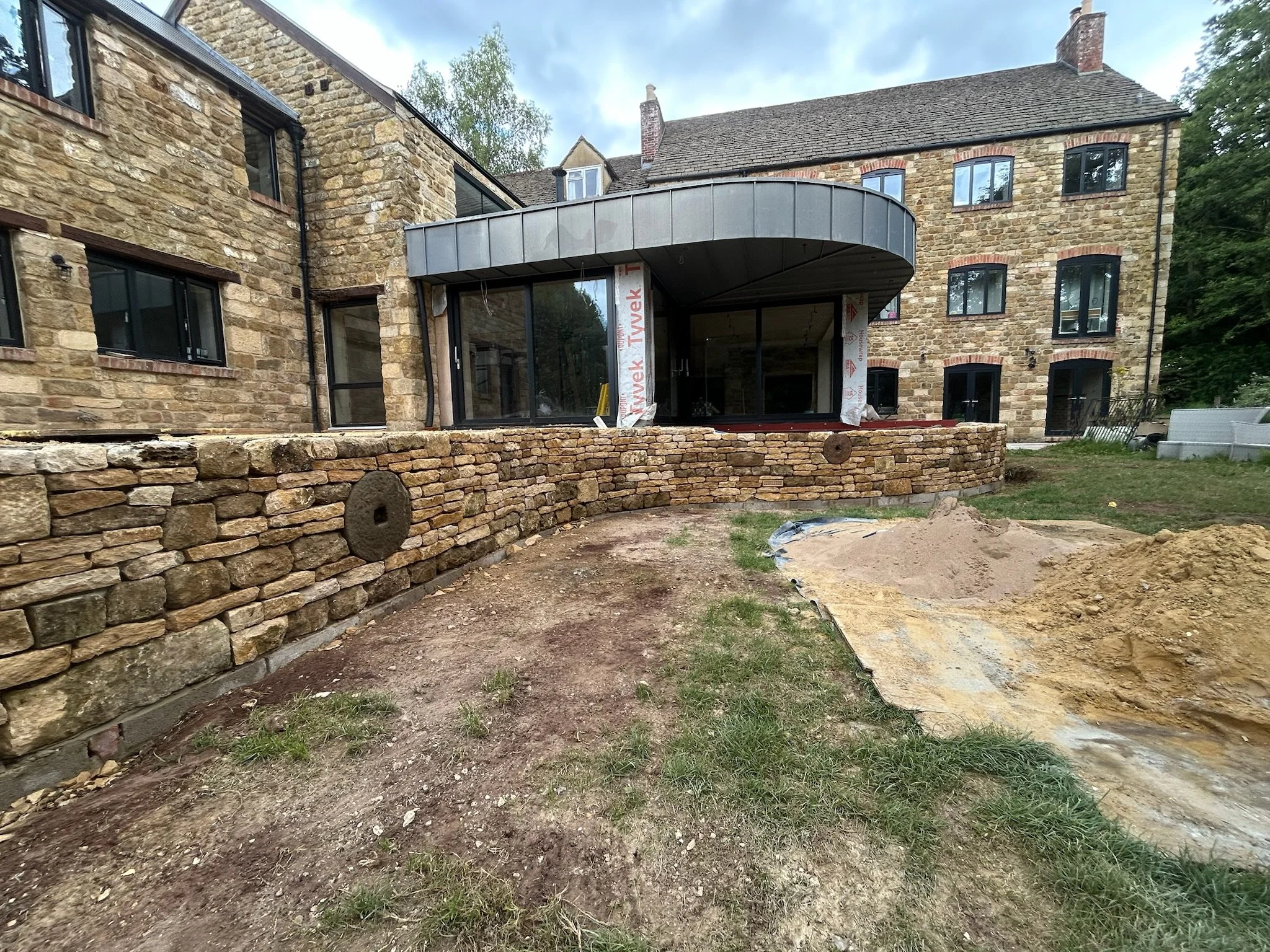 Construction site of a stone wall in front of a multi-story brick building with large windows and a curved balcony or roof extension, with dirt and sand piles in the foreground.