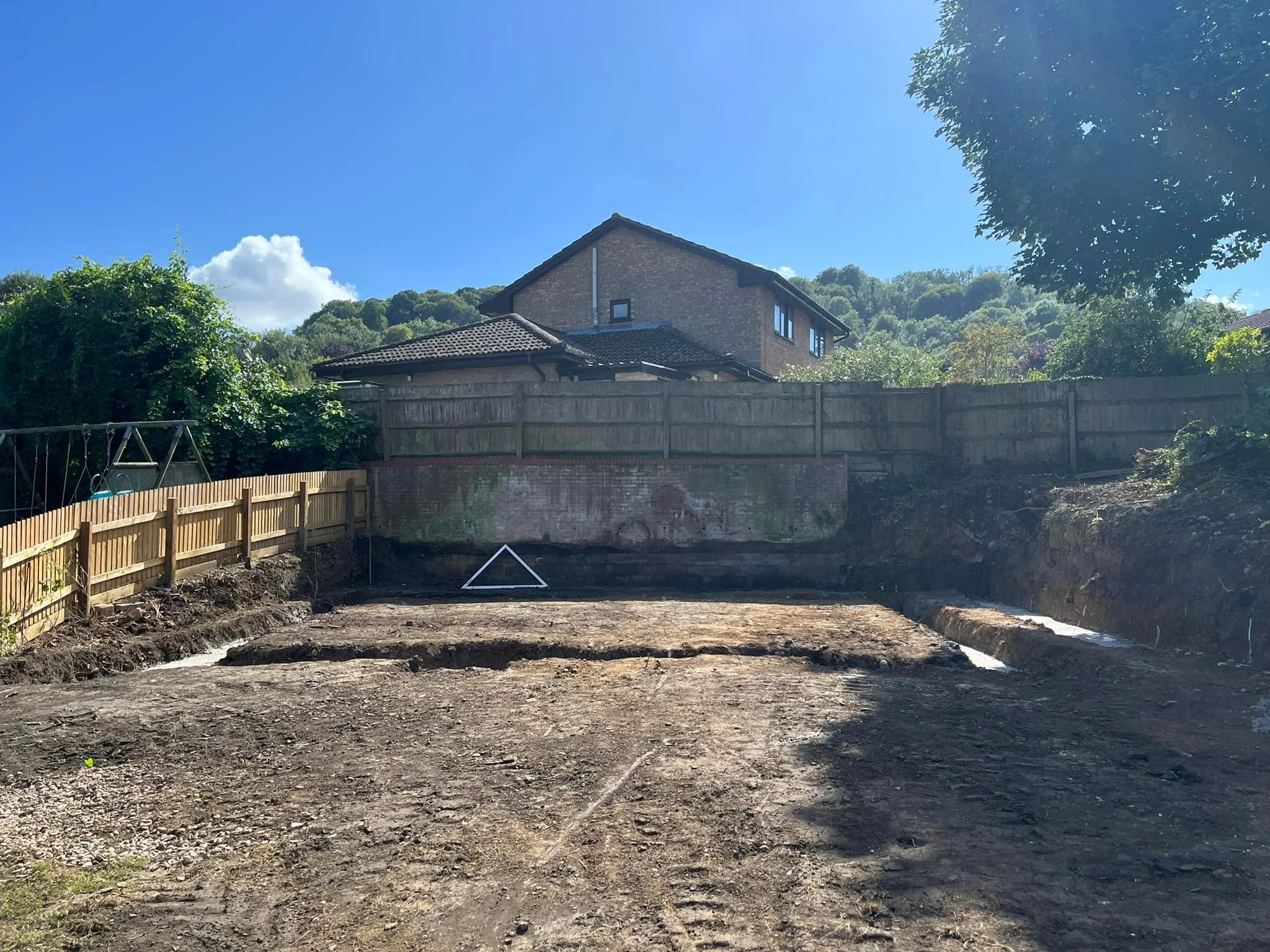 A residential backyard under construction, with a cleared dirt area, wooden fences, and houses with greenery in the background under a clear blue sky.
