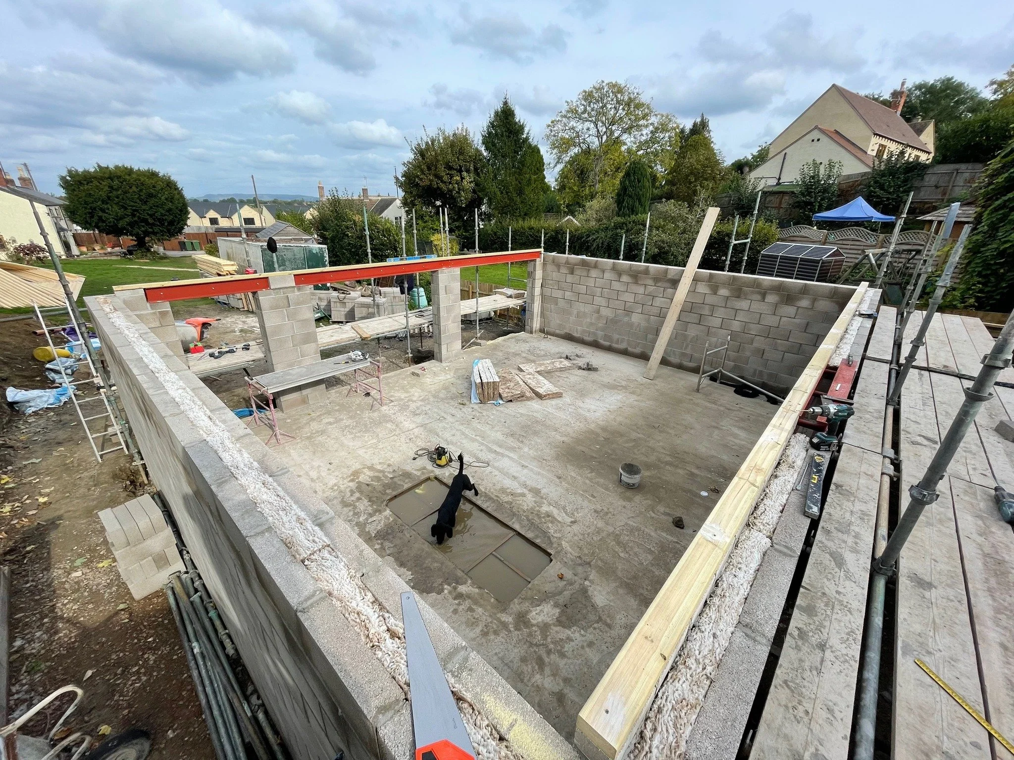 Construction site of a house foundation with cinder block walls partially built, construction tools, scaffolding, and building materials visible, in a residential neighborhood with trees and houses in the background.