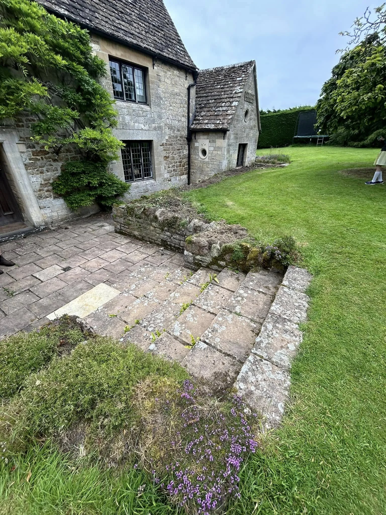 A stone house with a sloped roof, surrounded by green grass and bushes, with a small stone pathway and steps in the yard.