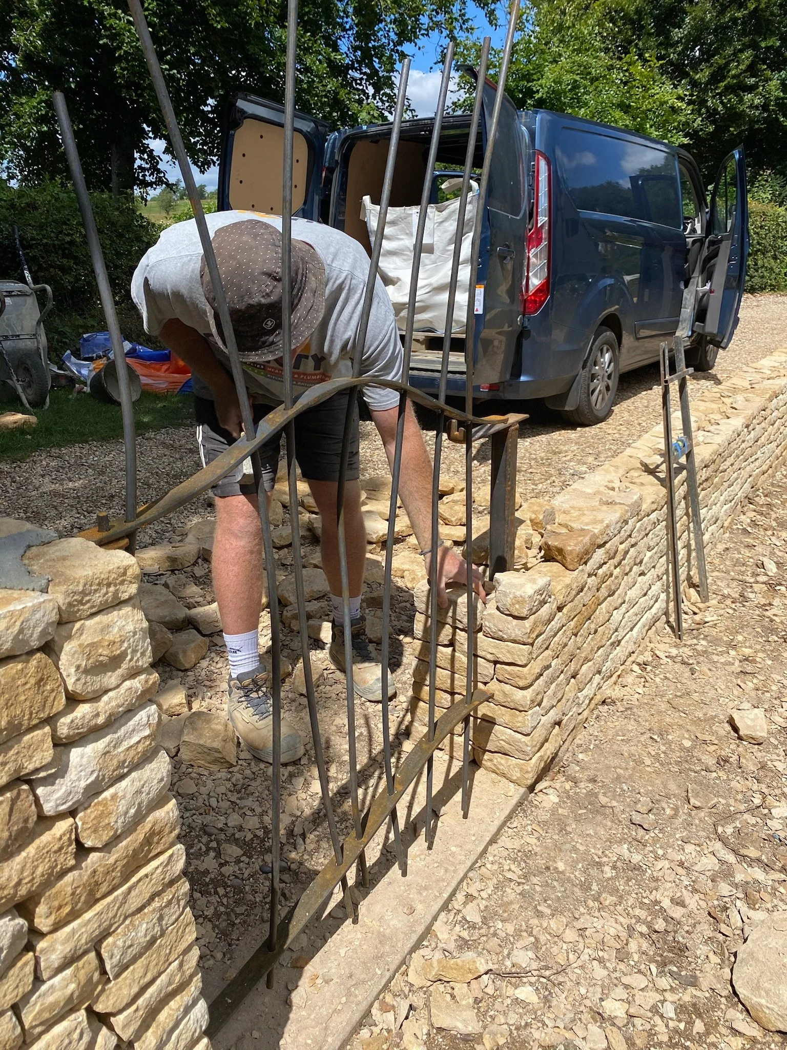 A man working on a low stone wall with a metal fence in front of it, near a blue van with open doors, outdoors with trees and a gravel ground.