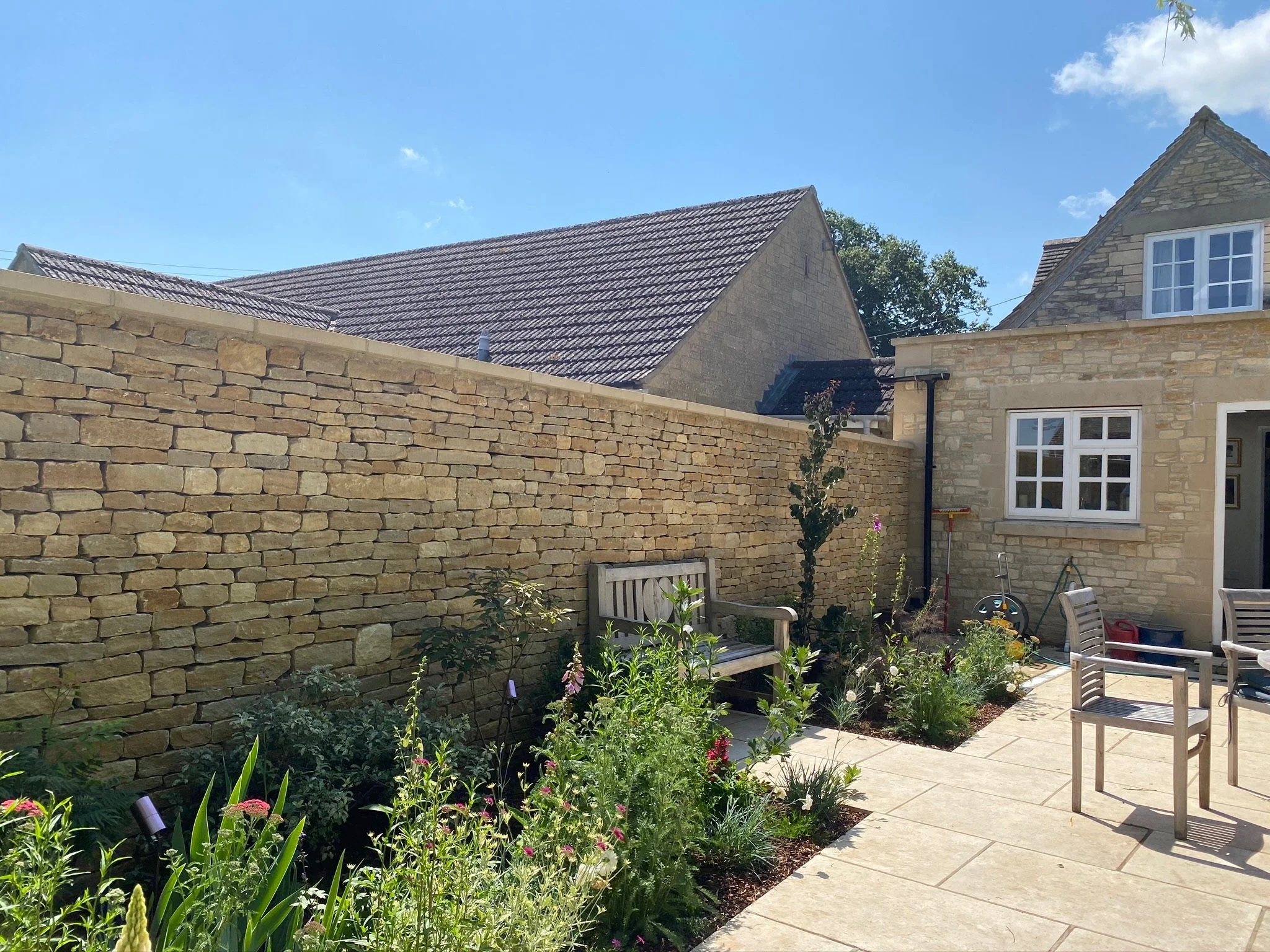 A backyard garden patio with potted plants and outdoor seating, enclosed by a stone wall and neighboring stone houses under a bright blue sky.