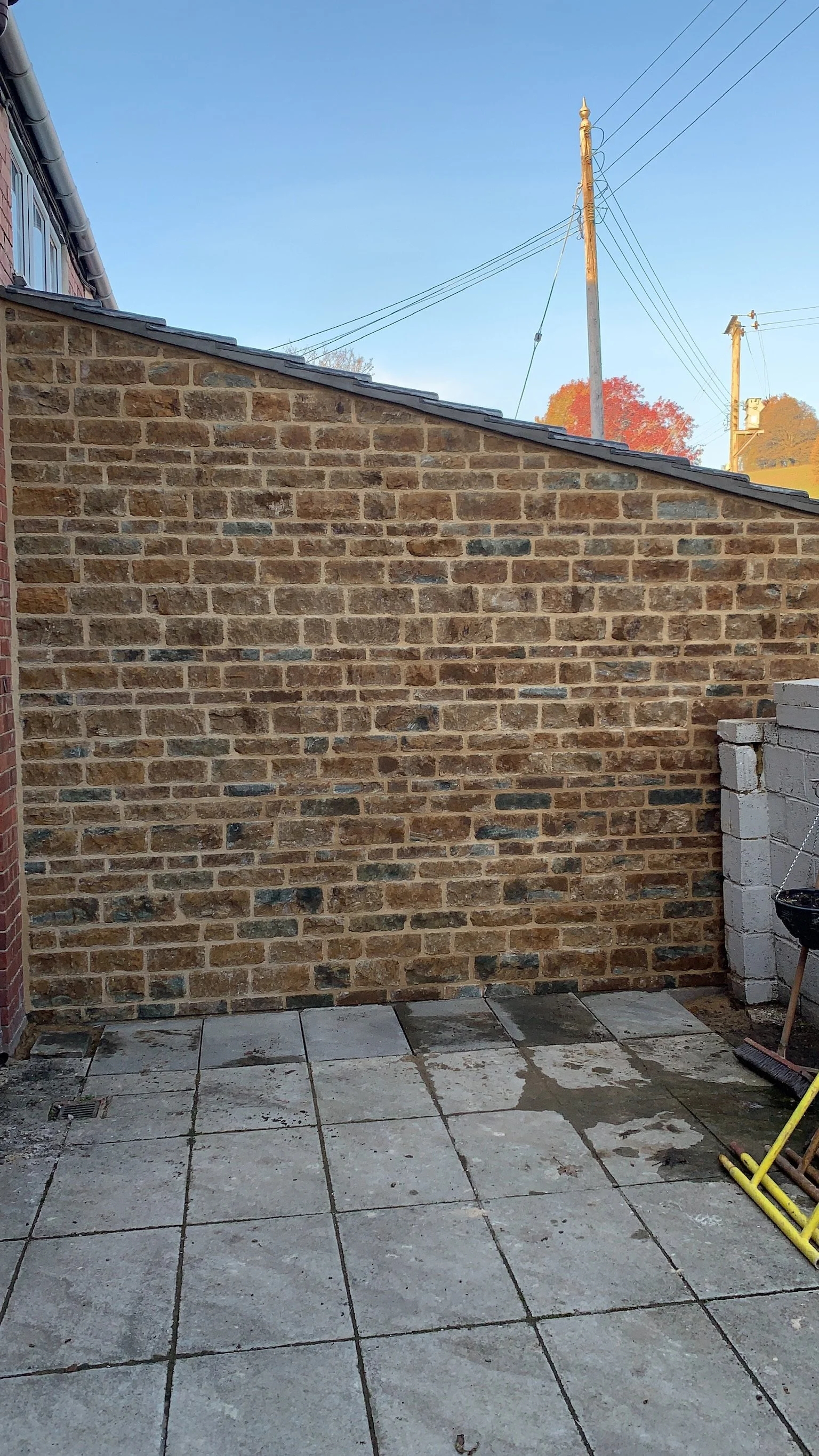 Exterior view of a brick wall with a sloped roof, some construction materials, and tools on a tiled patio, under a clear blue sky.