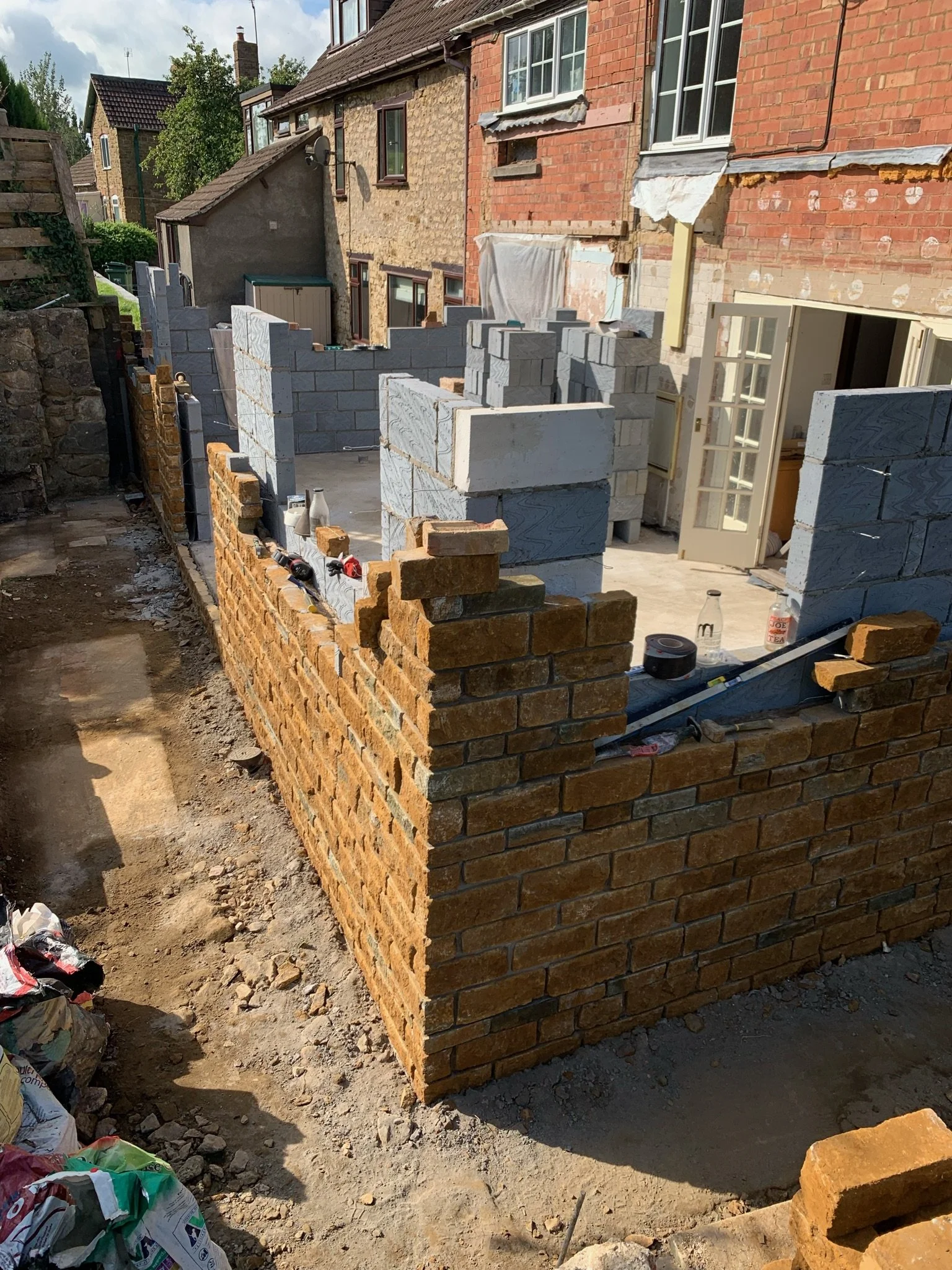 Construction site with a brick wall partially built in front of a residential building, with construction tools and materials scattered around.