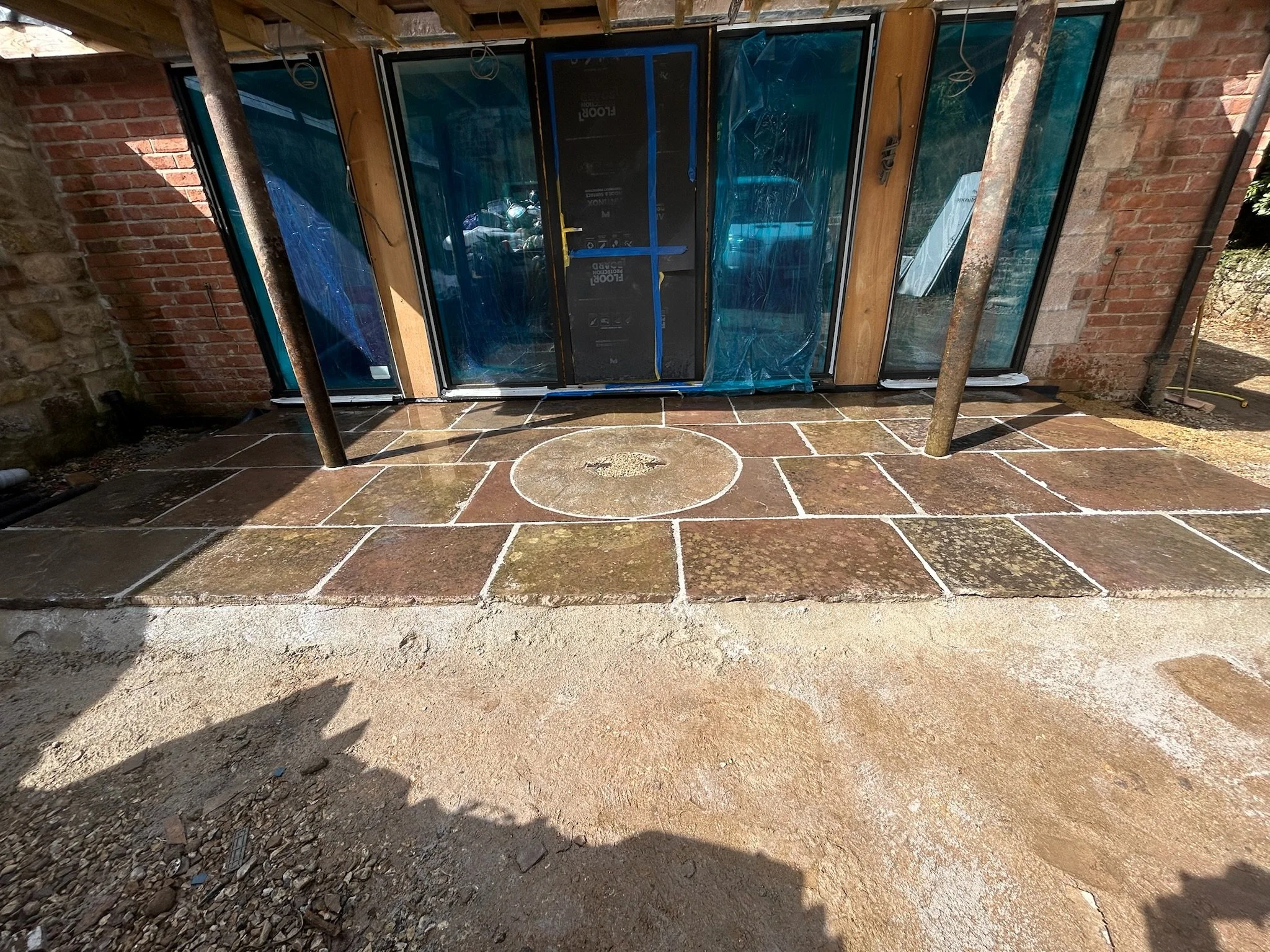 Construction site with newly laid stone patio in front of a brick building with glass windows. Support poles are in place, and construction materials are visible inside.