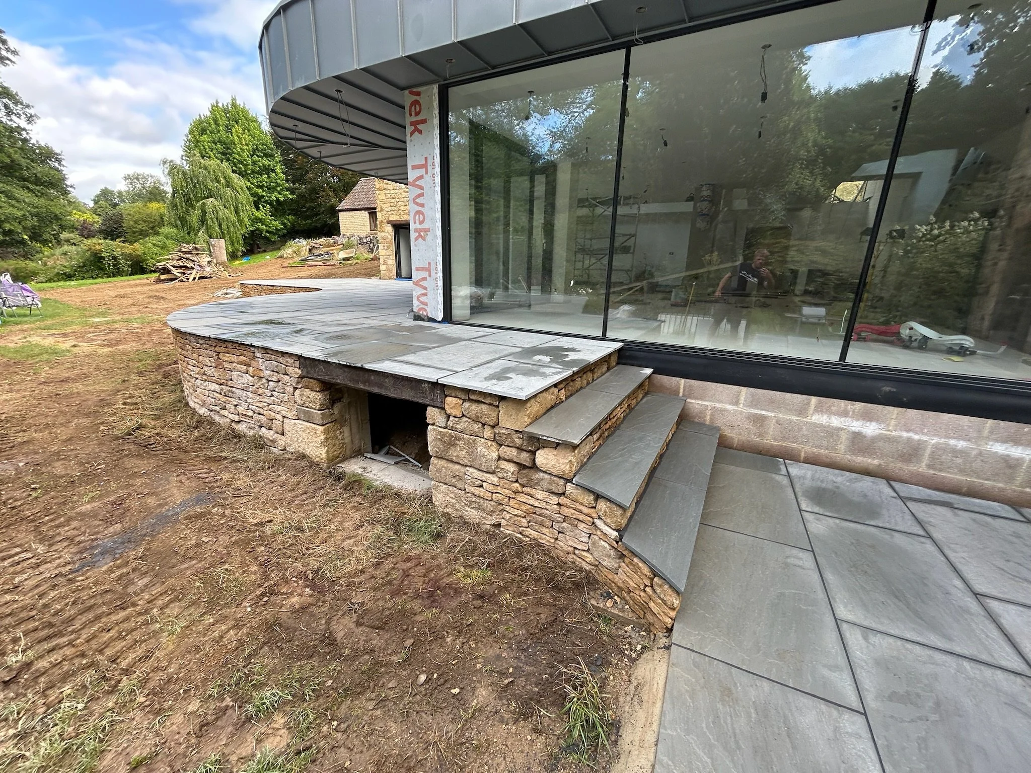 Under construction stone and slate steps beside a large glass window on a house with a curved exterior wall, with garden area and trees in the background.