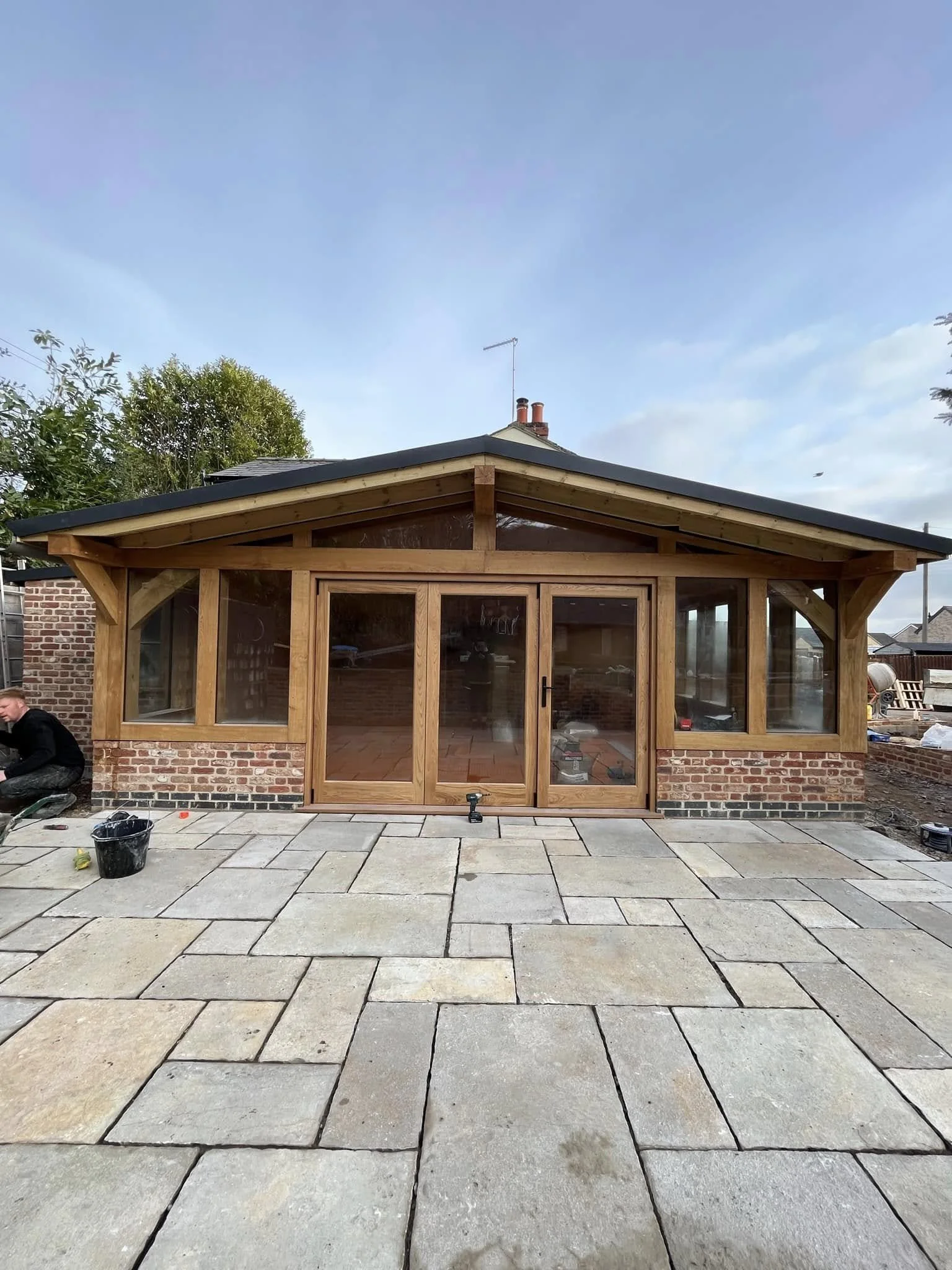 A newly constructed wooden extension with glass doors and large windows on a brick house, set on a stone-paved patio with construction tools and a worker on the left.