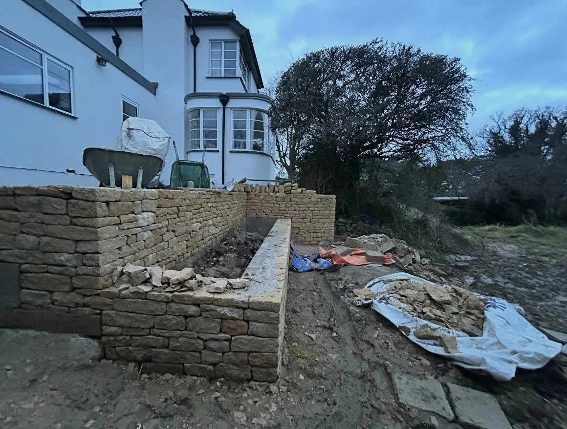 Construction site with stone wall and building in background, construction materials and tools scattered around.