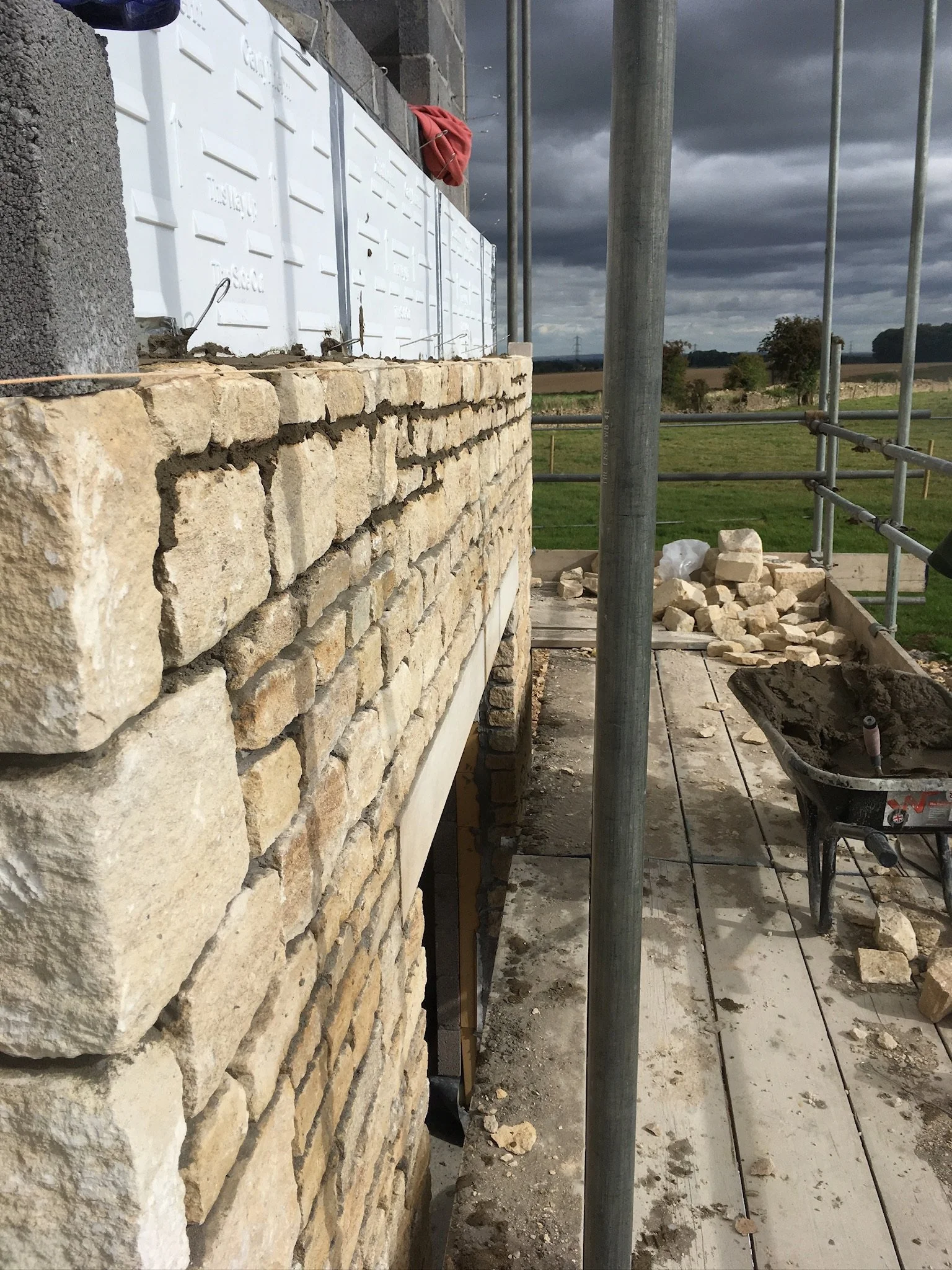 Construction site showing a stone wall being built with scaffolding and construction materials near a field with cloudy sky in the background.