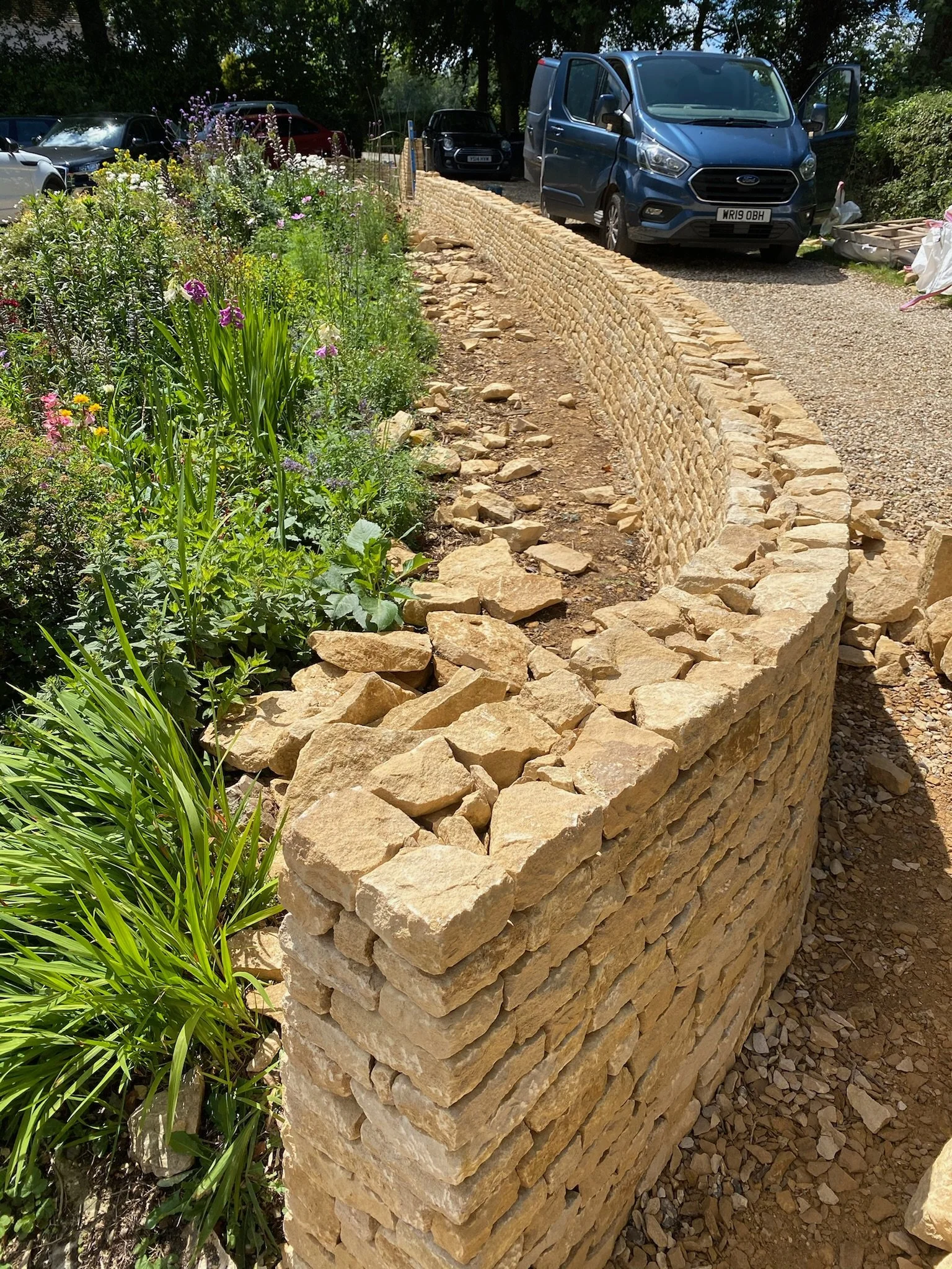 A curved dry-stone wall made of beige stones separates a garden with colorful green plants and flowers from a parking area with several parked cars and a blue van.