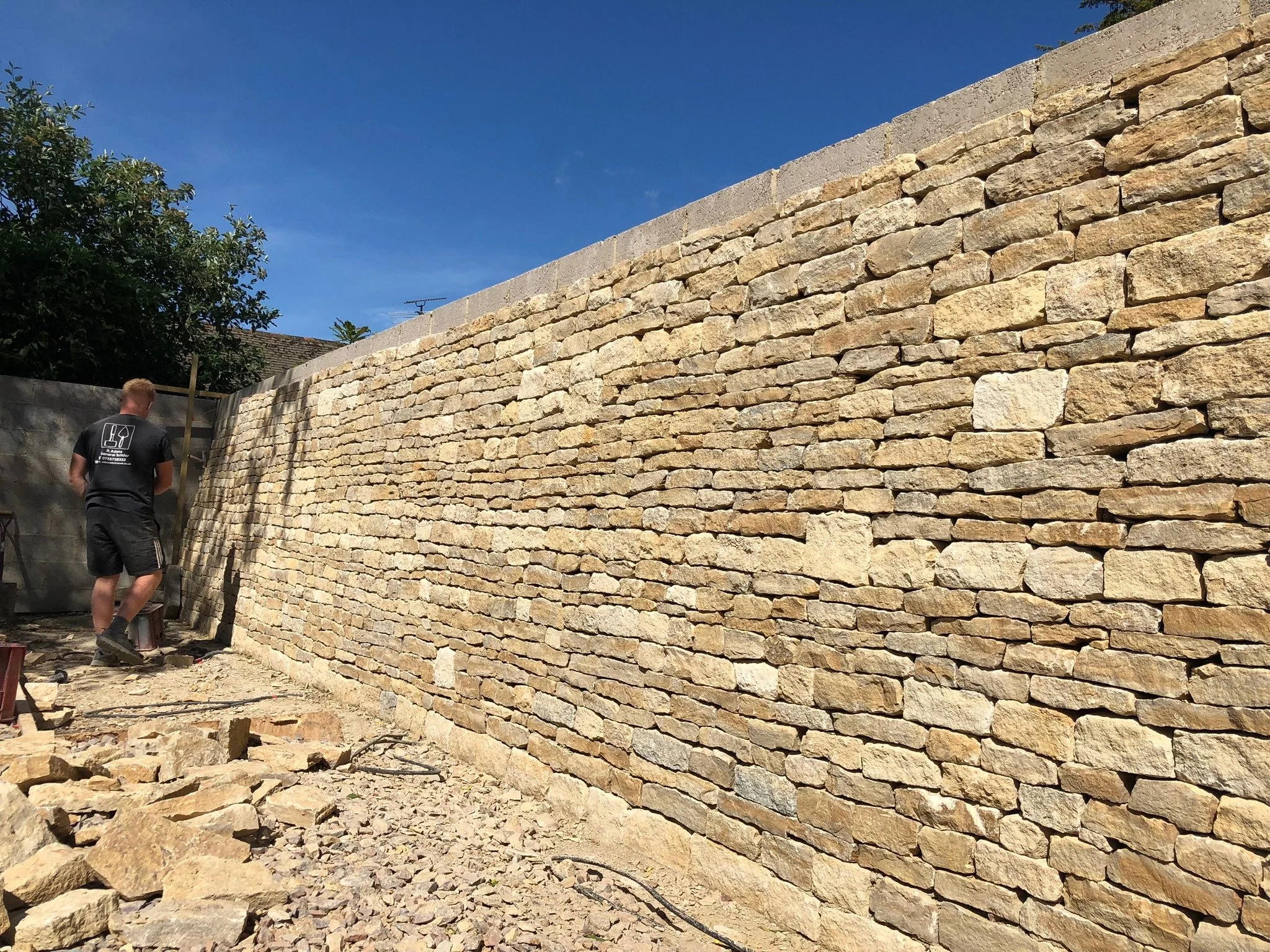 A worker building a stone wall outdoors under a clear blue sky.