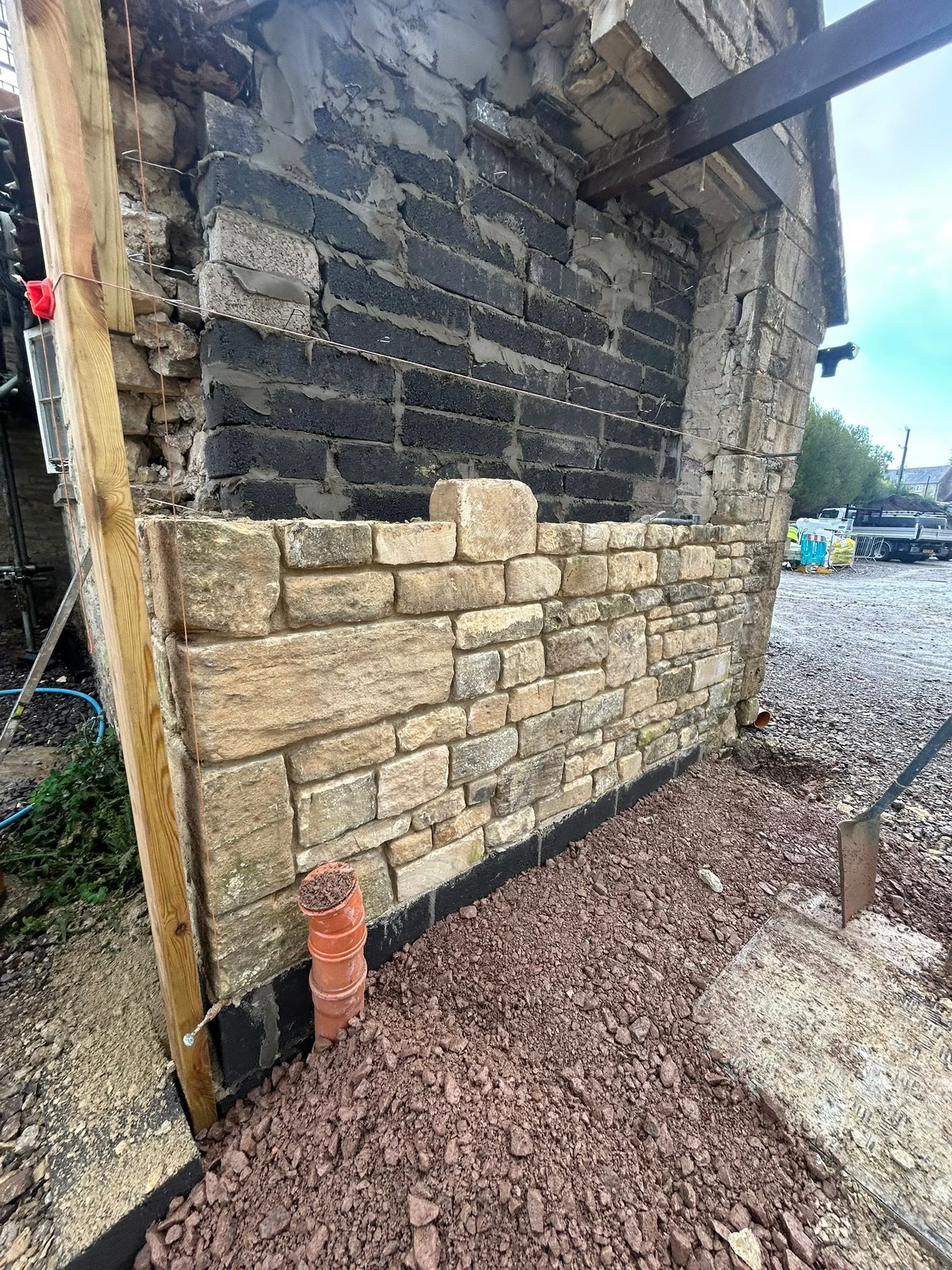 Construction site showing a brick wall in progress with black and beige stones, a wooden support beam, and a dirt ground with a shovel.