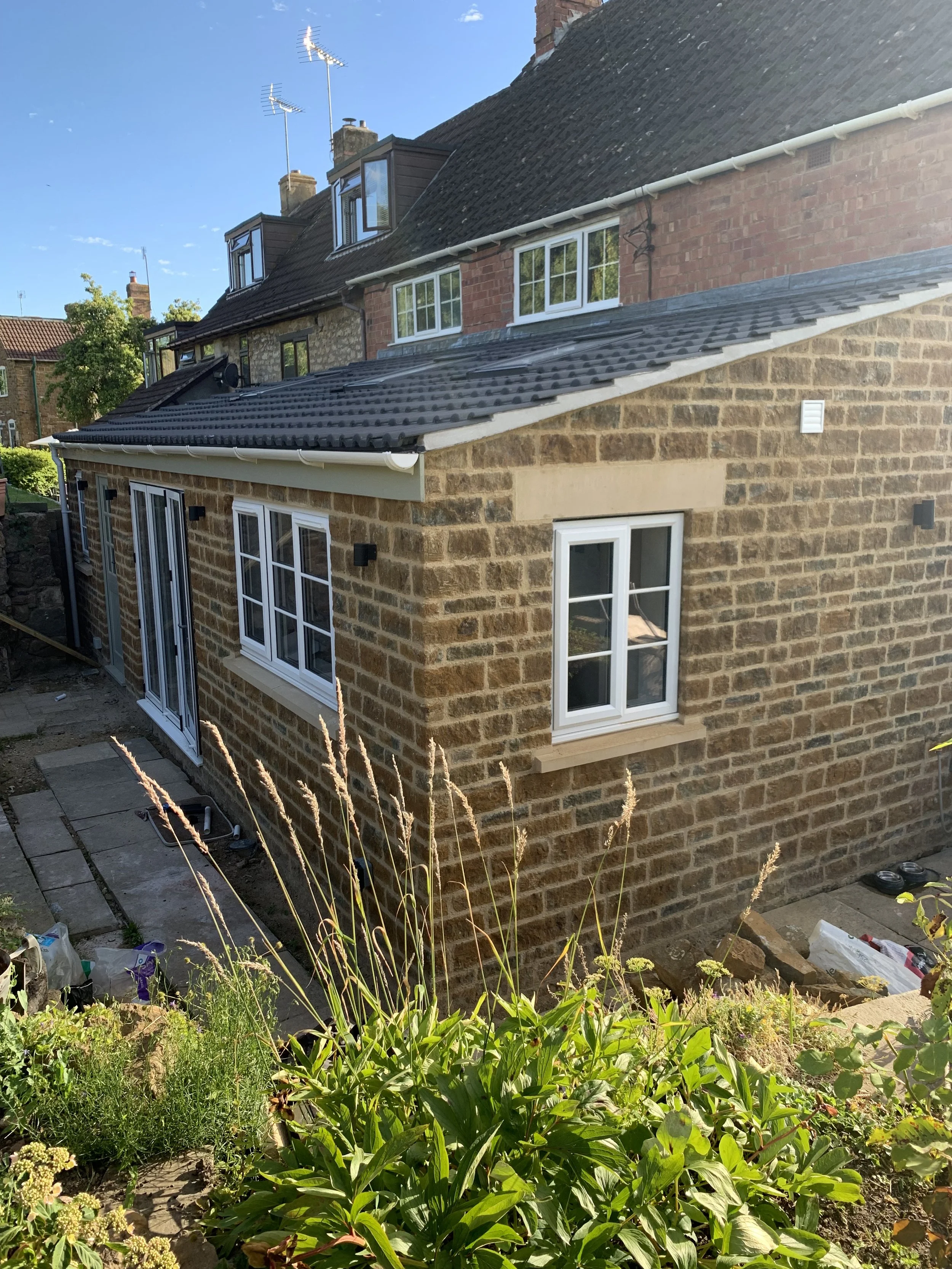 A brick house with white-framed windows and a newly constructed extension, surrounded by a garden with green plants and tall grass, under a clear blue sky.