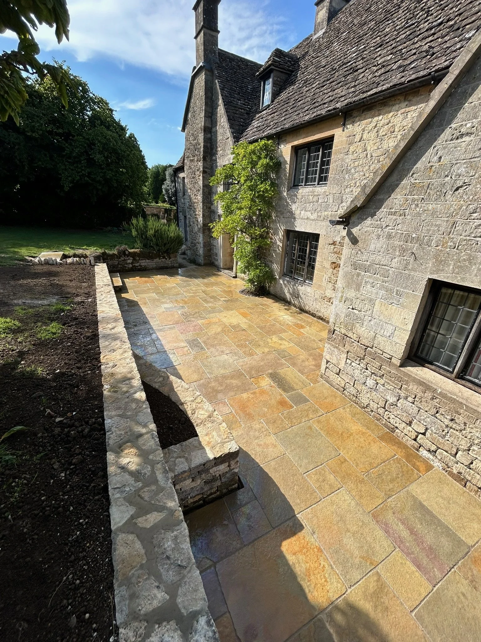 Stone patio in the backyard of a stone house with multiple windows and a small tree.