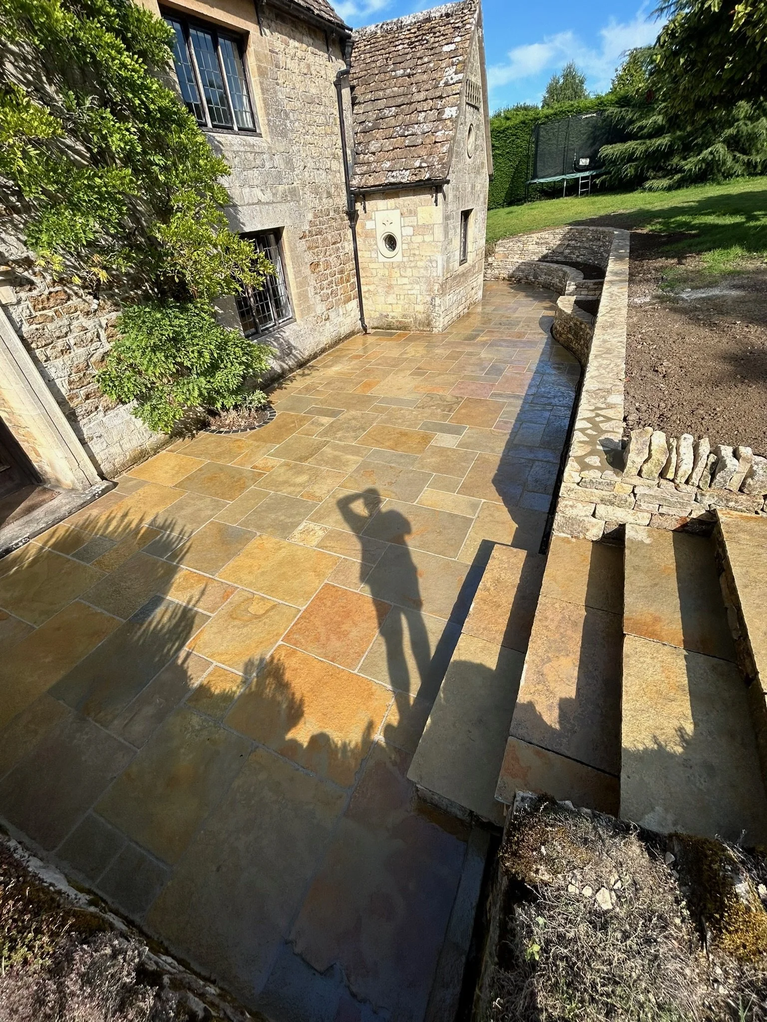 A sunlit stone patio outside a brick house with barred windows, with a person taking a photo whose shadow is visible on the patio.