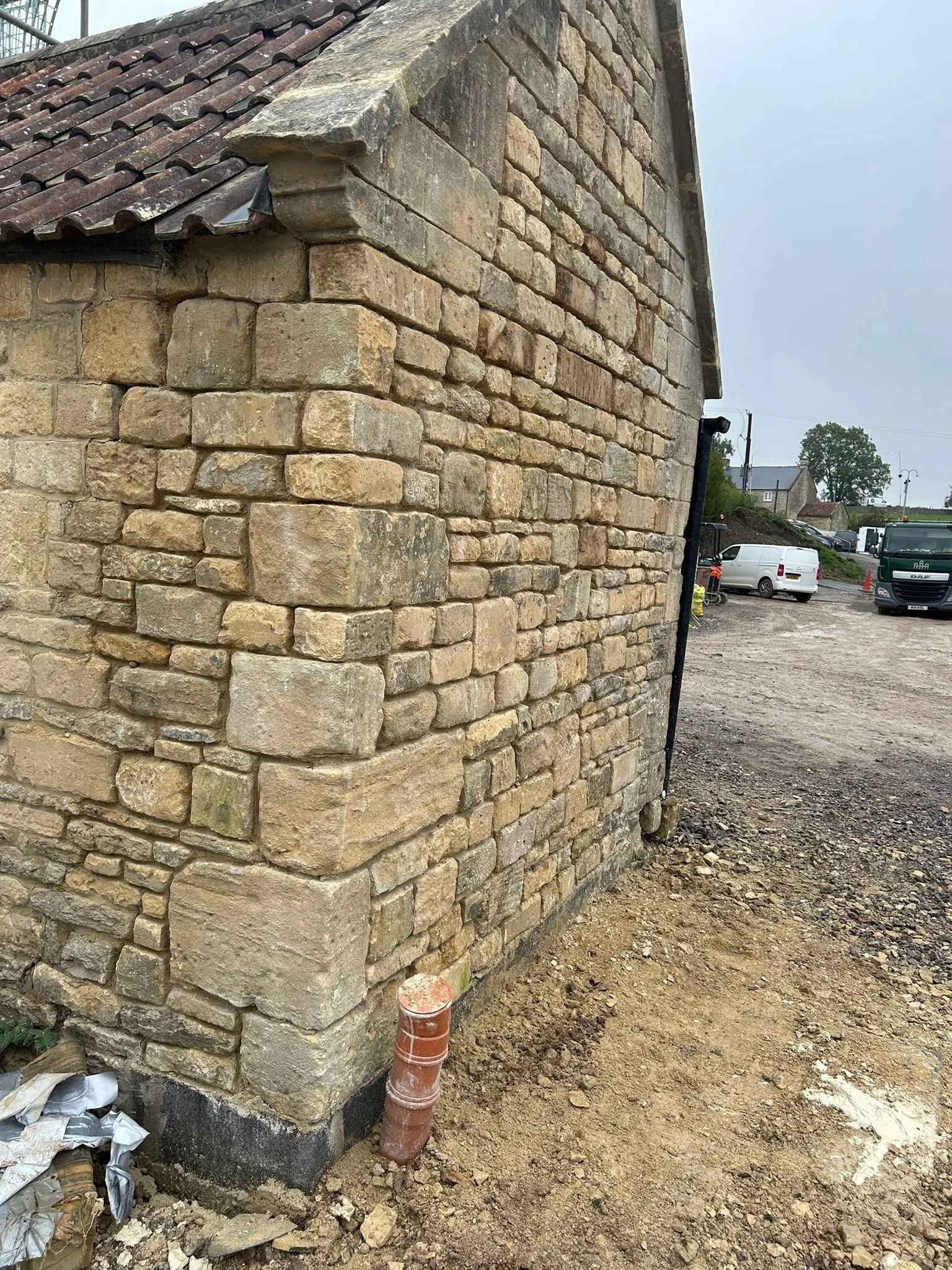 Close-up of a stone brick wall with new construction work at the base, including a metal pipe and construction materials on the ground, and a parking lot with vehicles in the background.