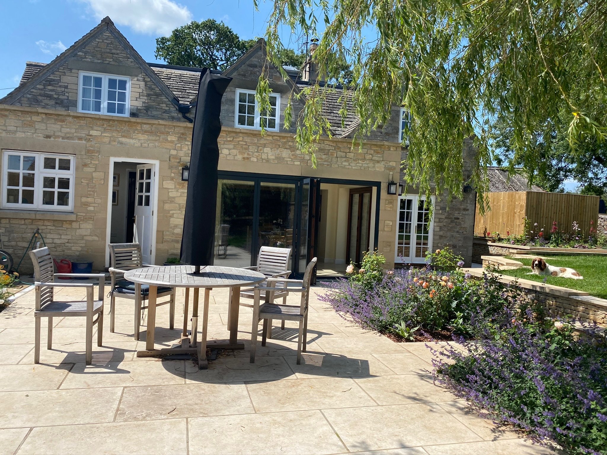 A backyard patio with a round wooden table and six chairs, with a black umbrella soaking up the sun. To the right, a dog is lying on the grass near a flower garden. The house in the background is made of stone with multiple white-trimmed windows and 