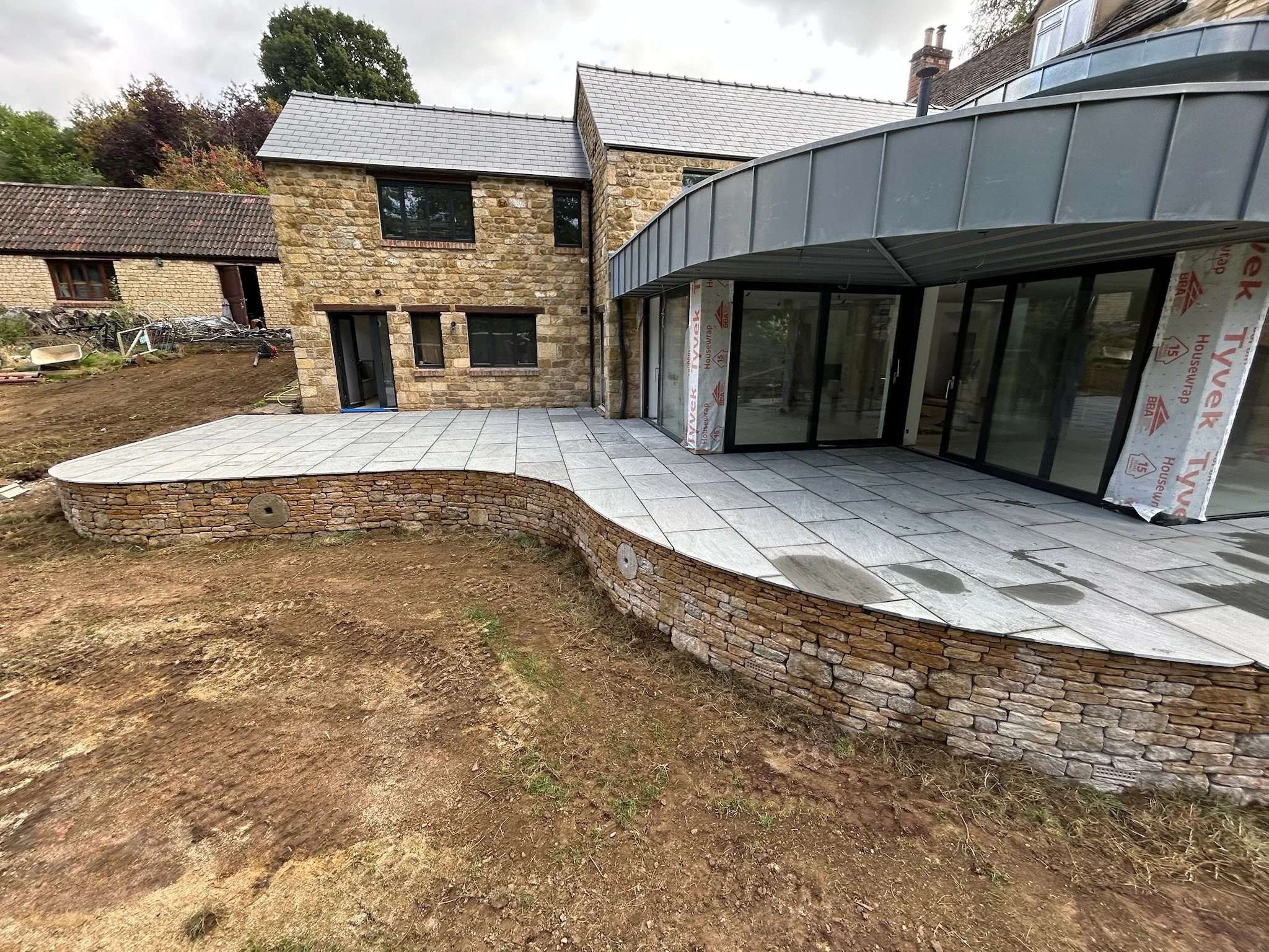 Newly constructed stone patio with a curved edge in the backyard of a stone house under construction, with large glass sliding doors and a metal roof extension.