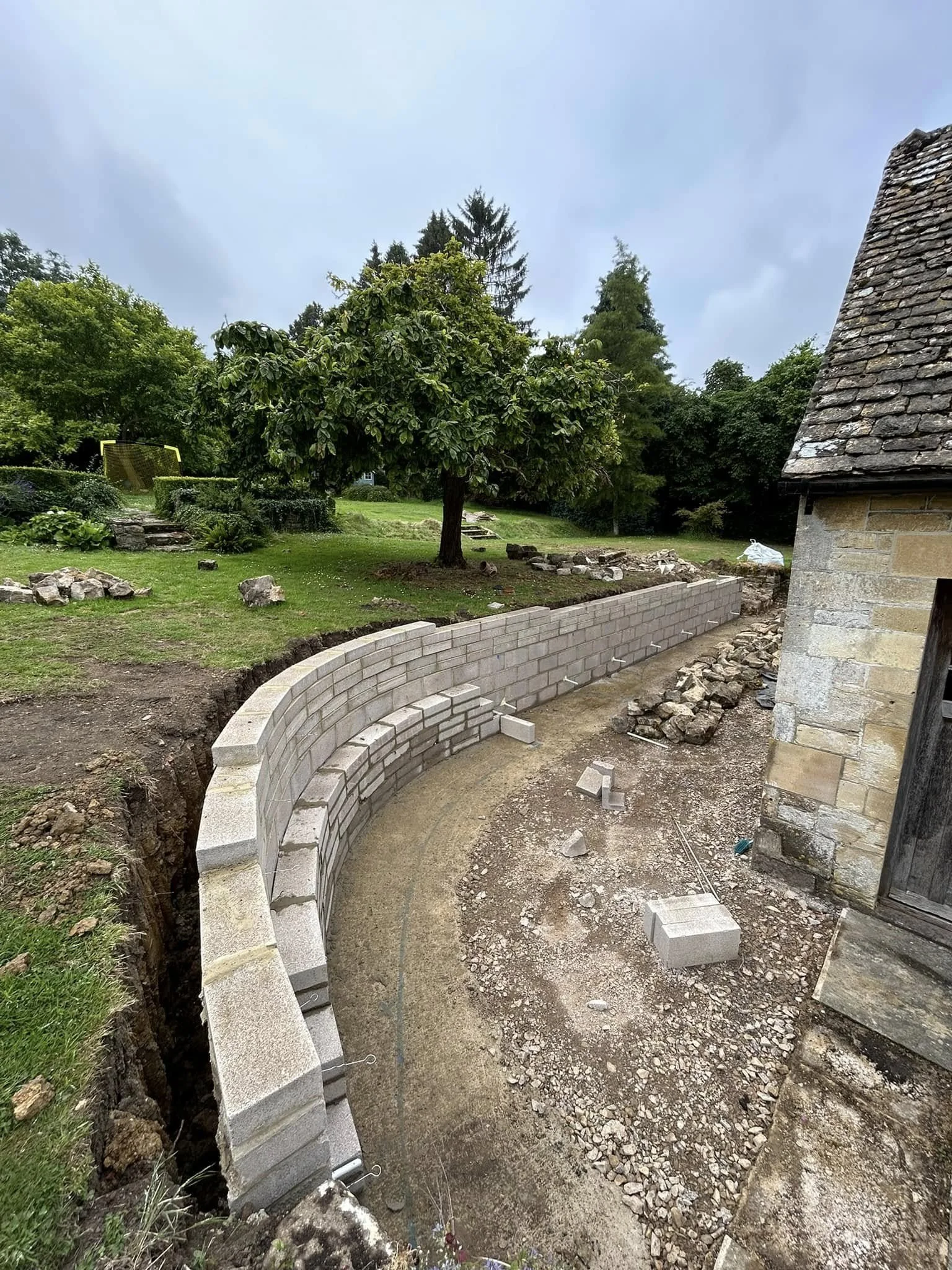 A curved stone wall construction in a backyard, with a green lawn, trees, and a stone building nearby.