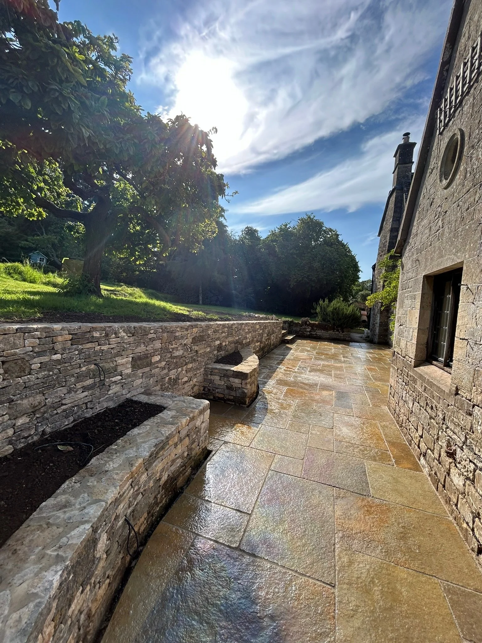 Sunlit stone patio next to a stone house with a chimney, bordered by a stone retaining wall with planter boxes, and a grassy area with a large tree under a partly cloudy sky.