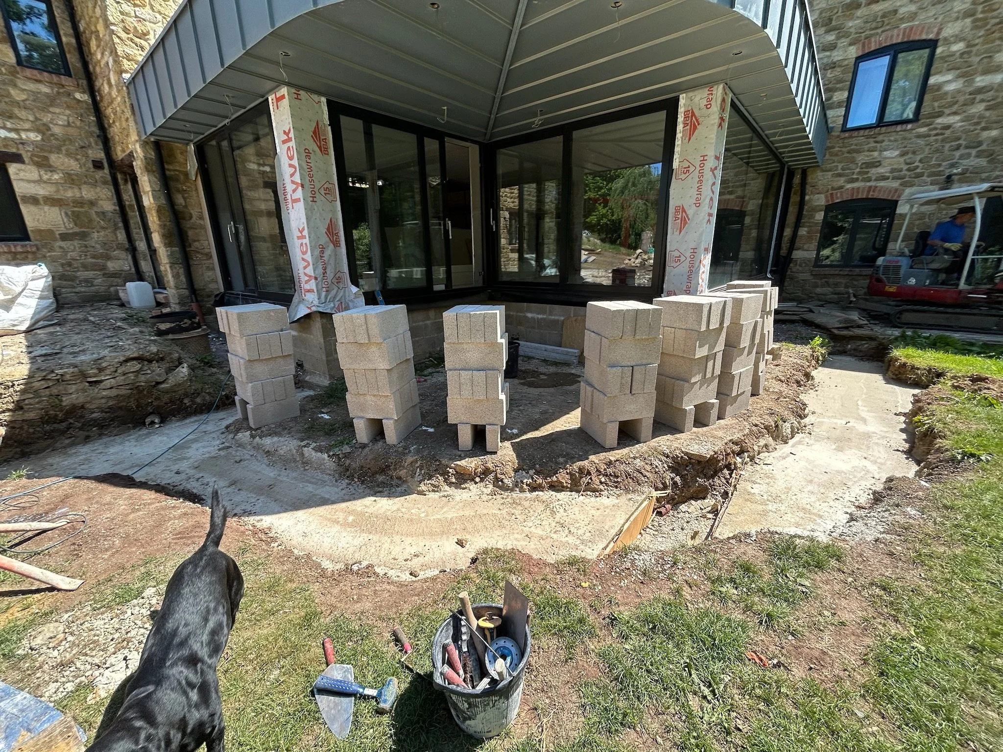 Construction site in the backyard of a house with a new glass addition, stacked cinder blocks, construction tools, and a dog in the foreground.