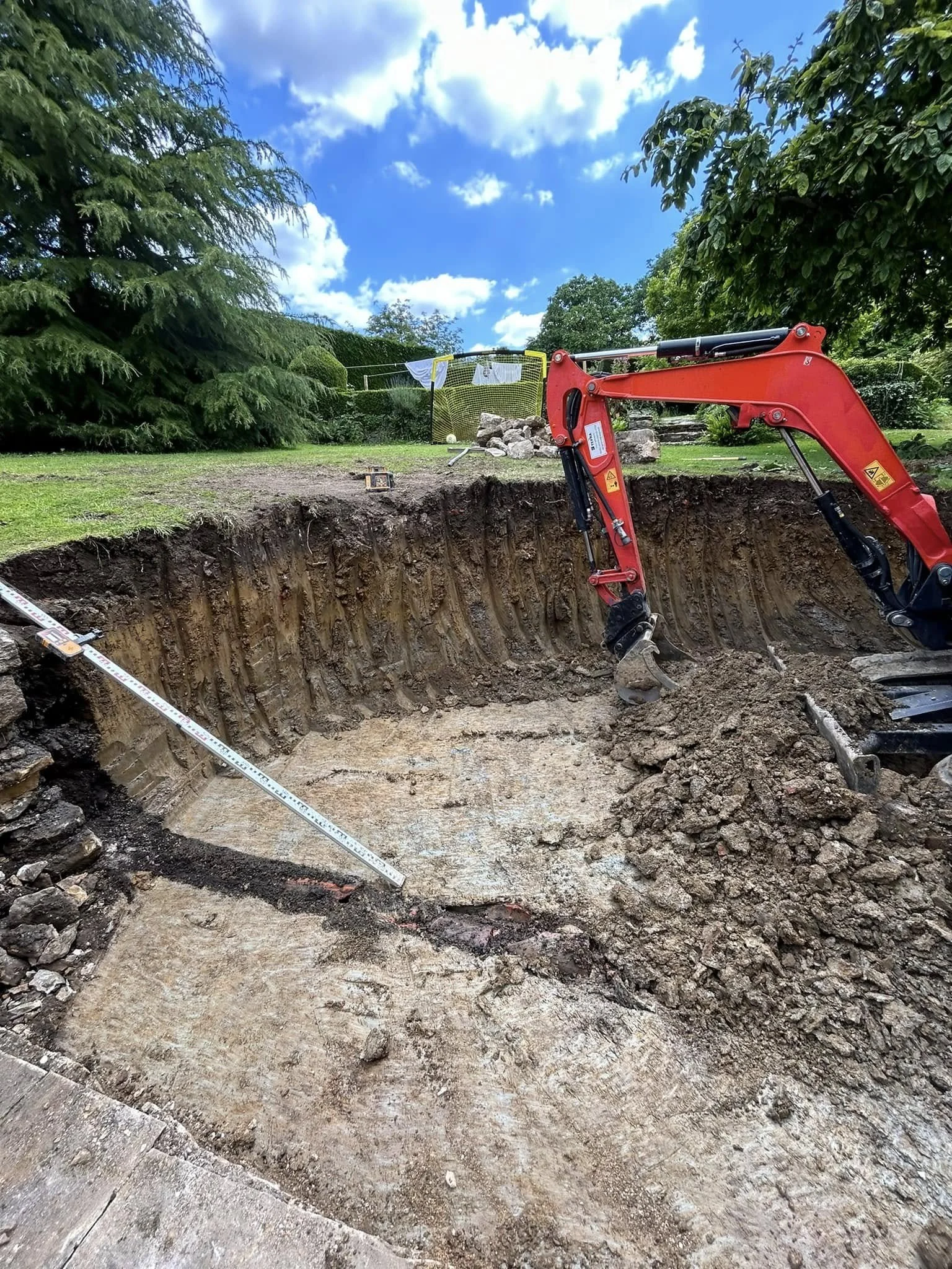 Construction site with an excavator digging a large hole in the ground, measuring tools visible, in a grassy area with trees, cloudy blue sky.
