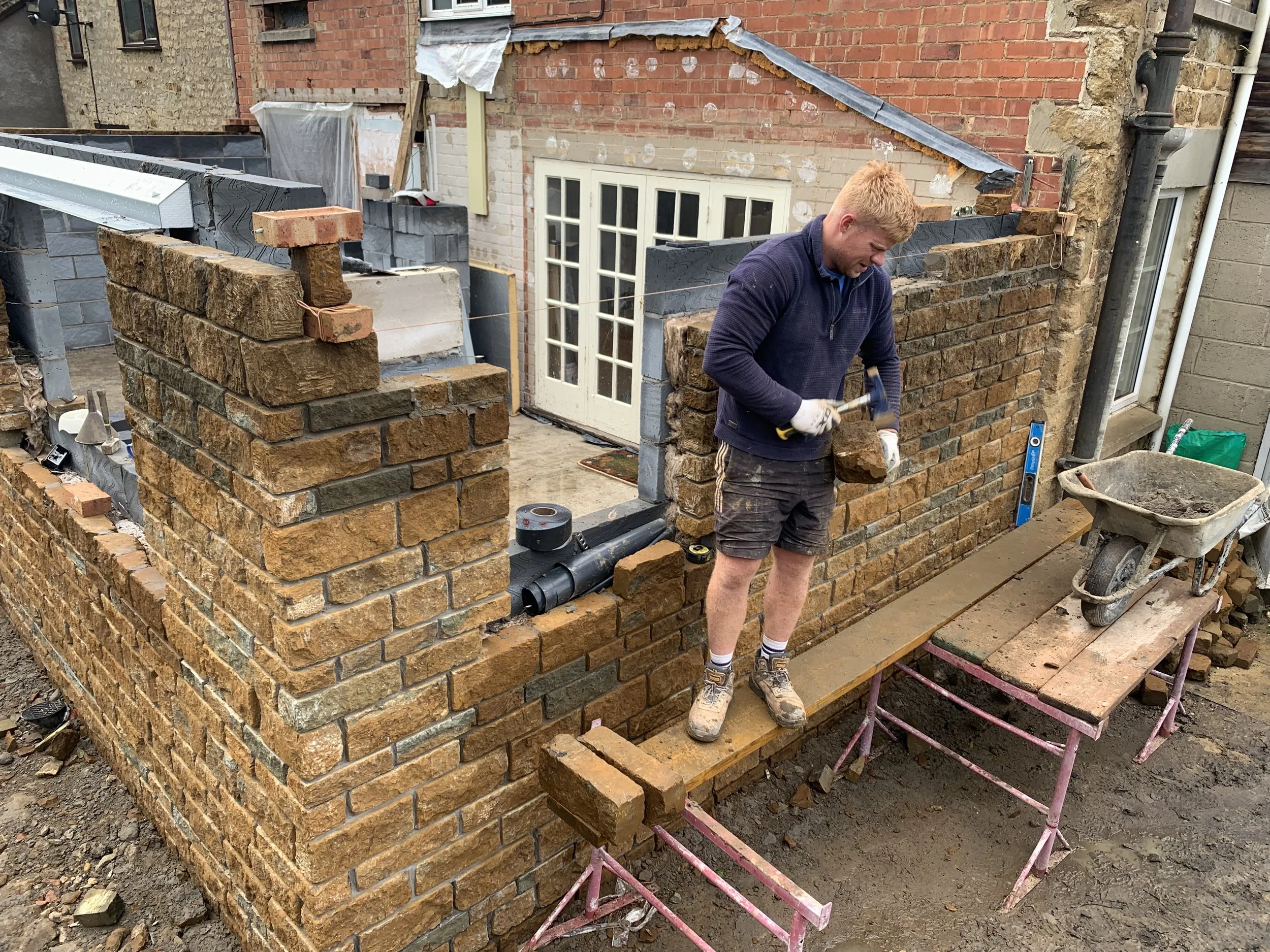A man is building a brick wall outdoors, using a hammer and a brick. He is standing on a wooden plank placed over pink scaffolding, with a wheelbarrow filled with mortar nearby. The wall is in progress with partially built sections, and construction 