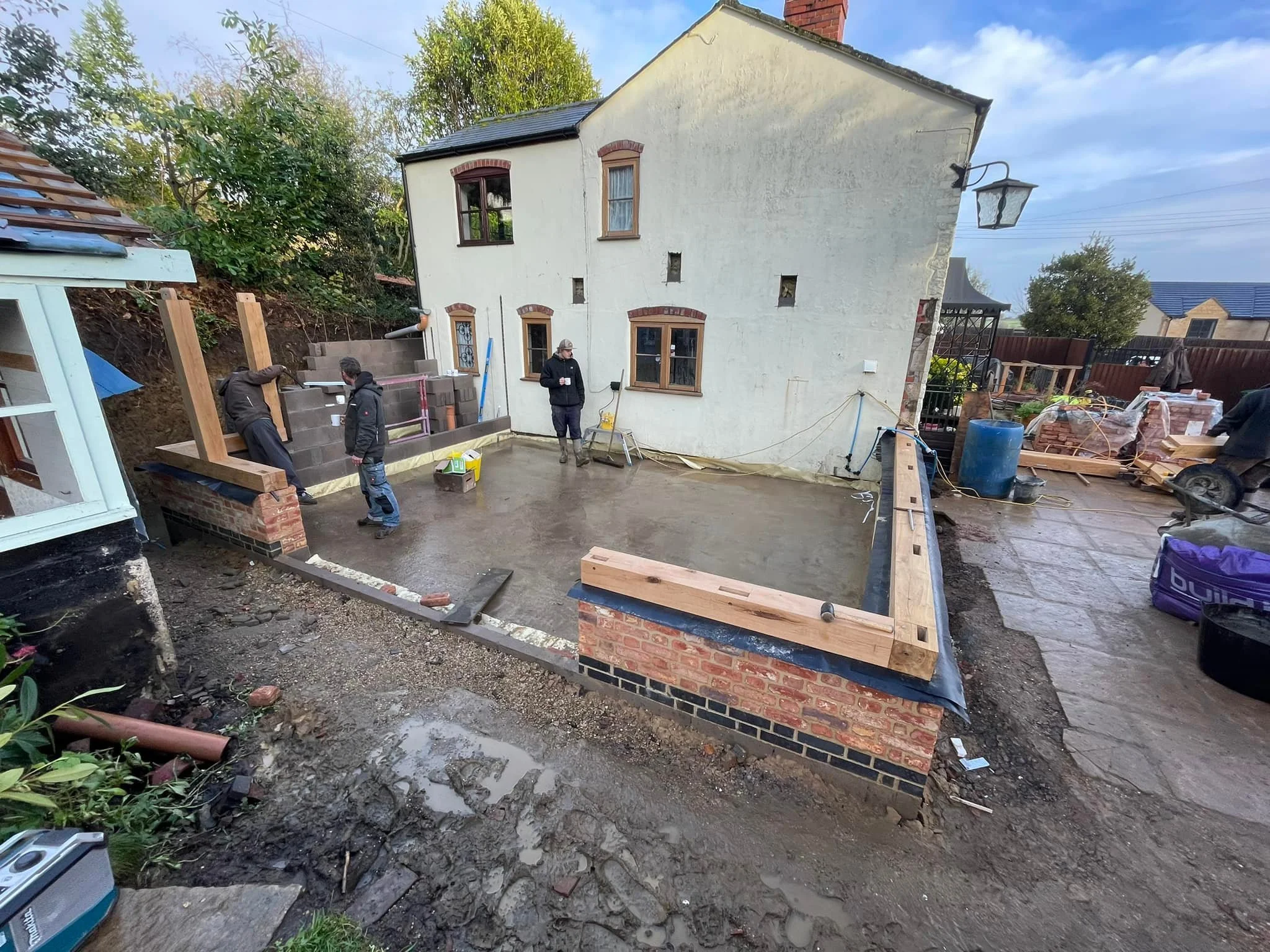 Construction workers inside a newly built brick patio poured with concrete, next to a white house with brown window frames, a blue rain barrel, and construction materials.