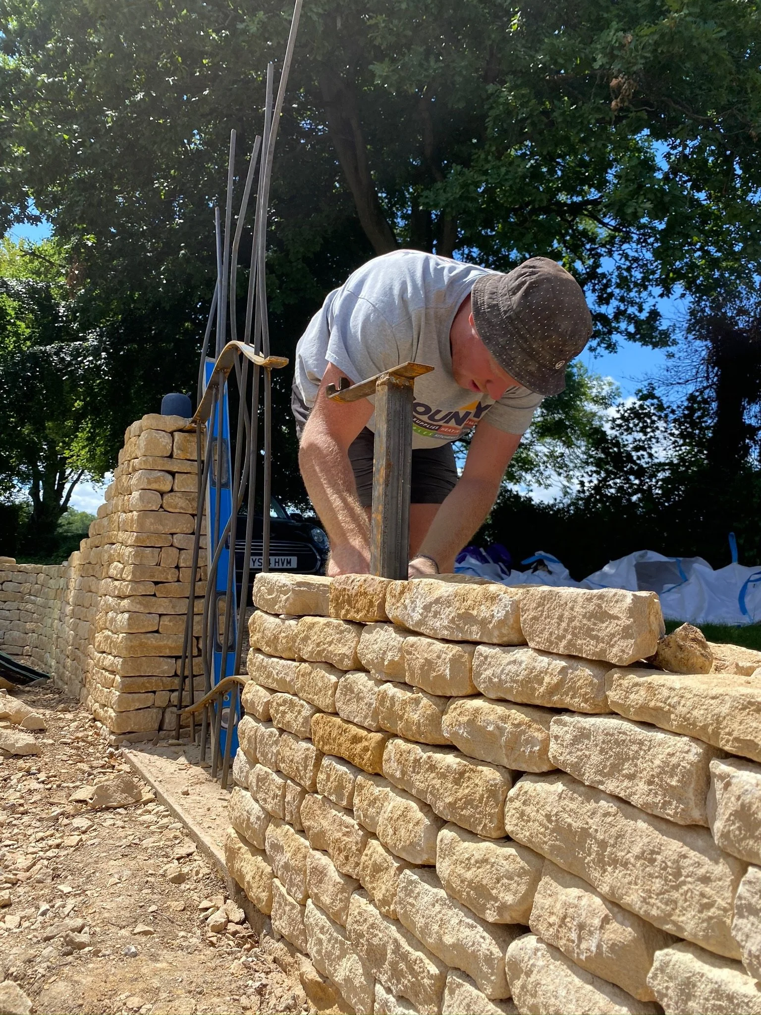 Man building a stone wall outdoors under a tree, using a hammer to set stones, with materials and tools nearby.
