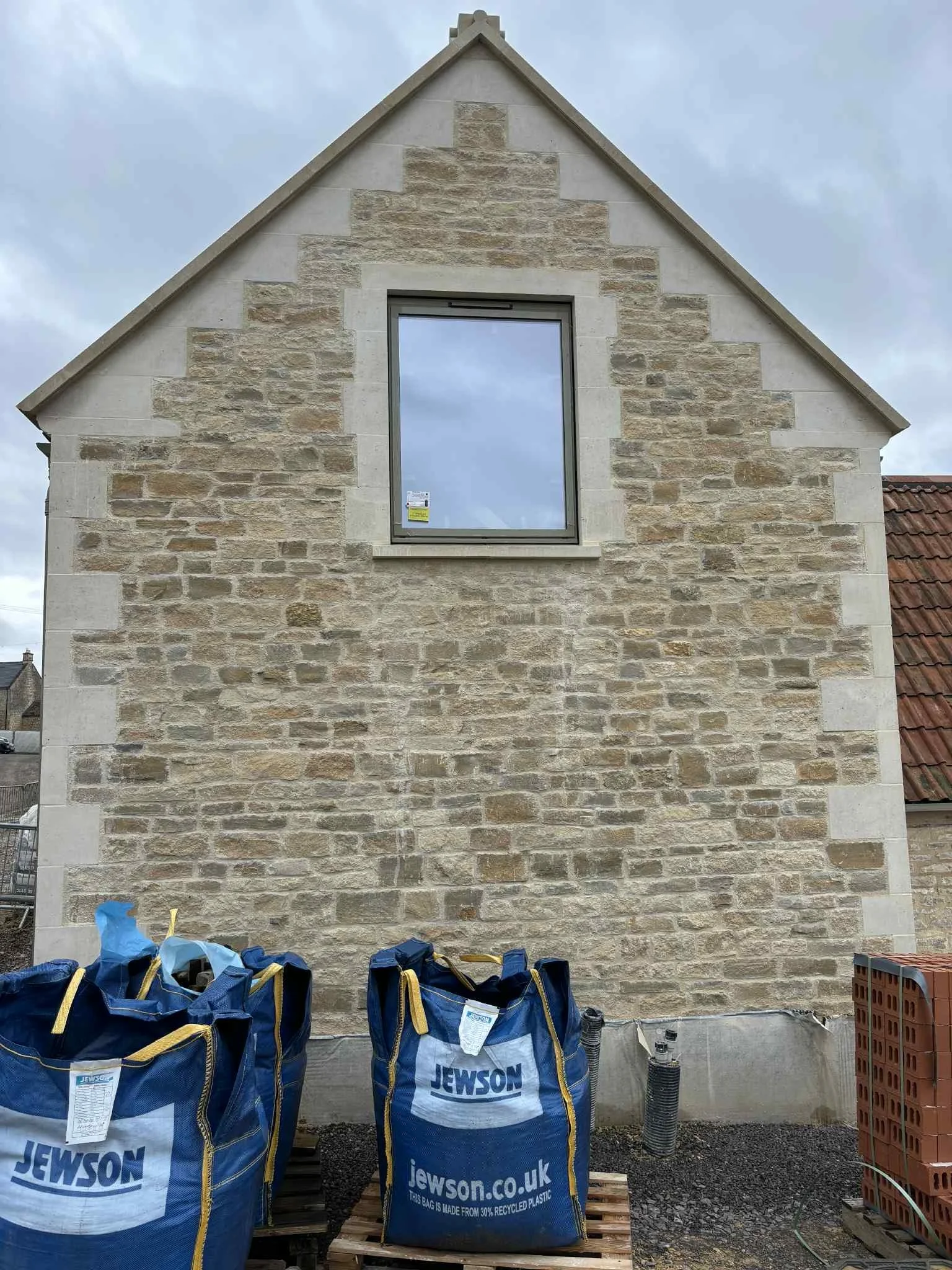 Construction site showing a stone house wall with a window, construction bags labeled 'Jewson' in the foreground, bricks, and building materials.
