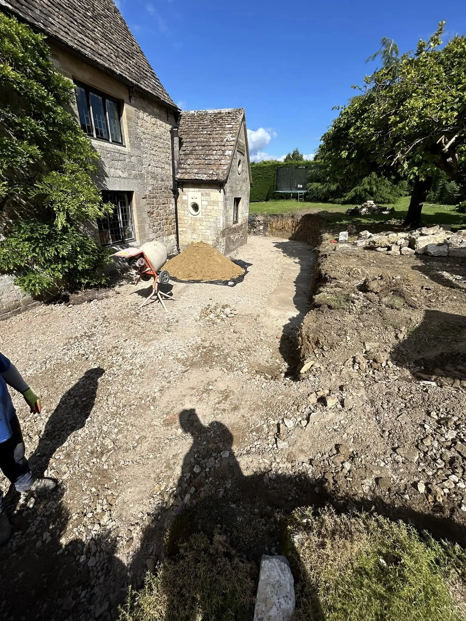 A construction site in a backyard with an old stone house. The ground is dug up, and gravel is spread around. There is a wheelbarrow, a pile of dirt, and some construction tools. Trees and a trampoline are visible in the background under a blue sky.