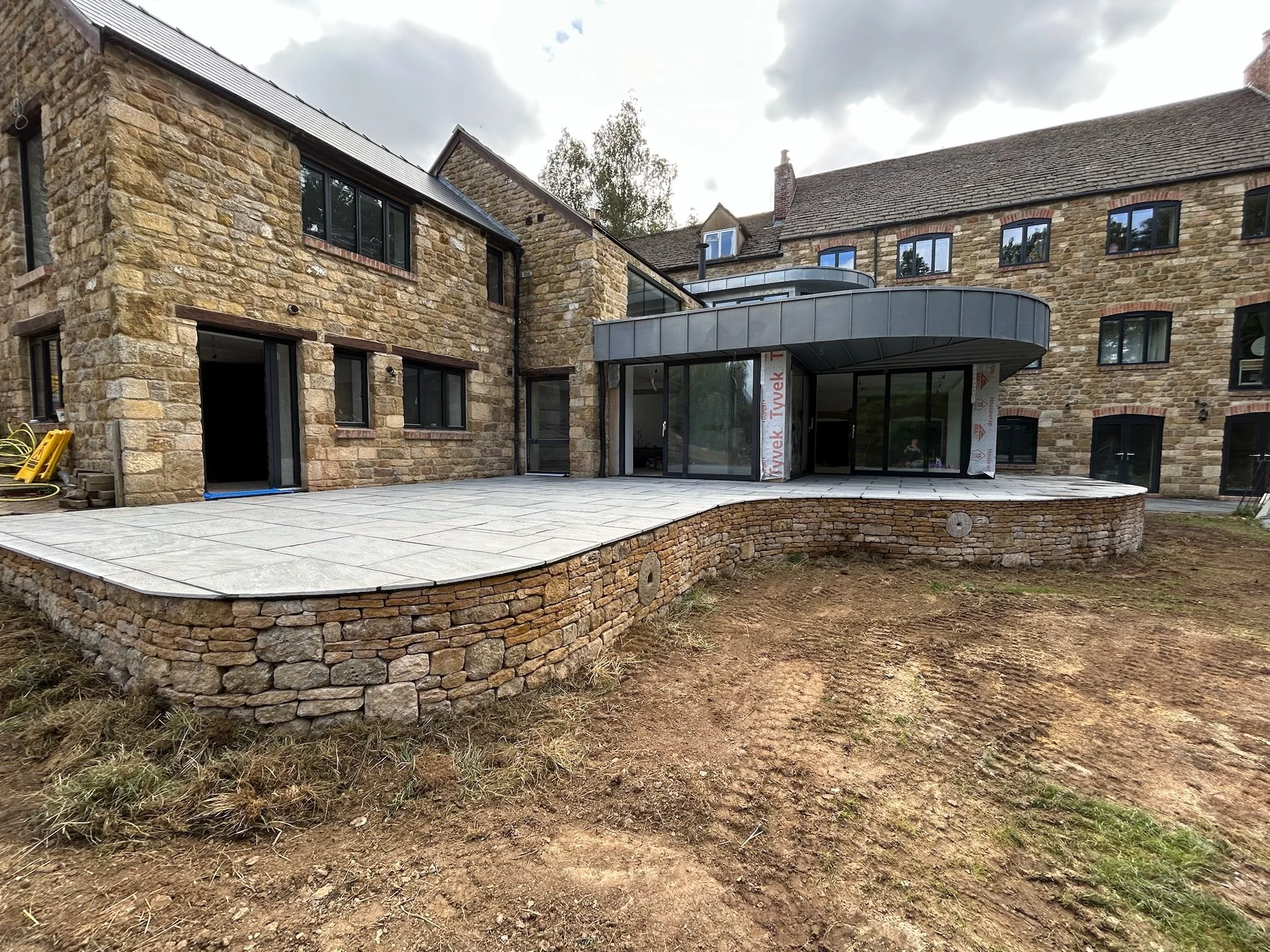 Construction site of a modern stone and brick building with a large patio area featuring large light gray tiles, and an area of bare dirt and grass, under an overcast sky.