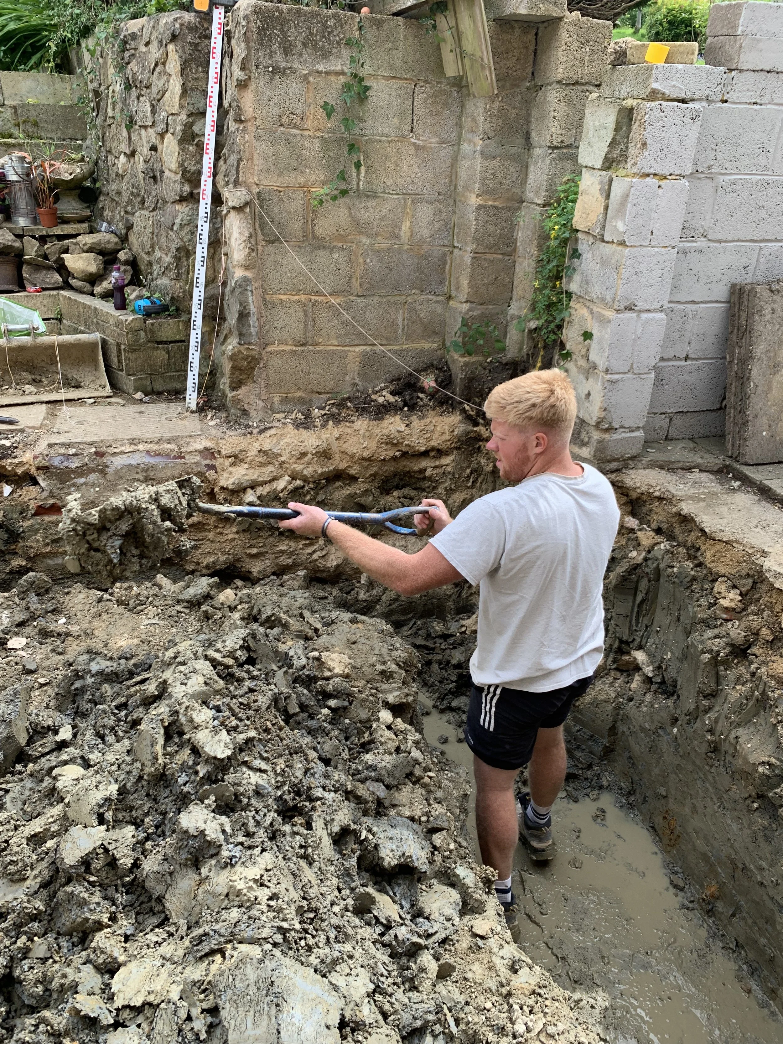 A person with blonde hair wearing a white t-shirt and black shorts is working in a deep trench with muddy water, using a power tool to break or remove concrete or dirt during excavation. Behind him are cinder block and stone walls.