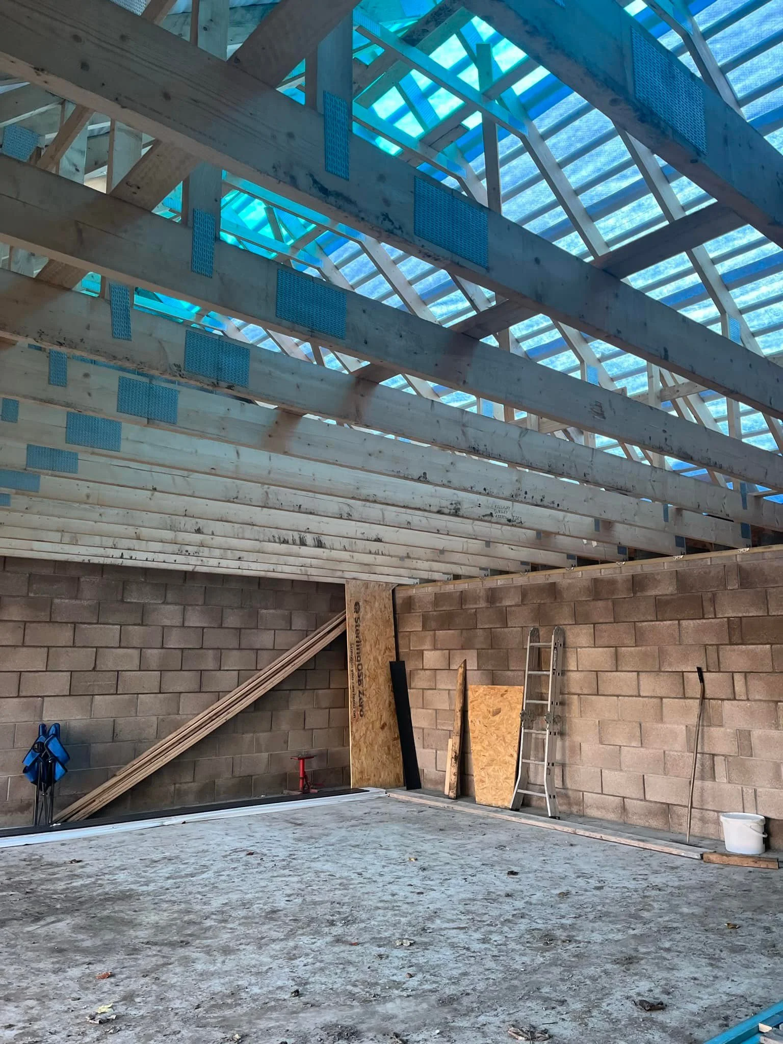 Interior of a construction site with a partially built roof, wooden beams, a ladder, stacked plywood, and a cinder block wall.