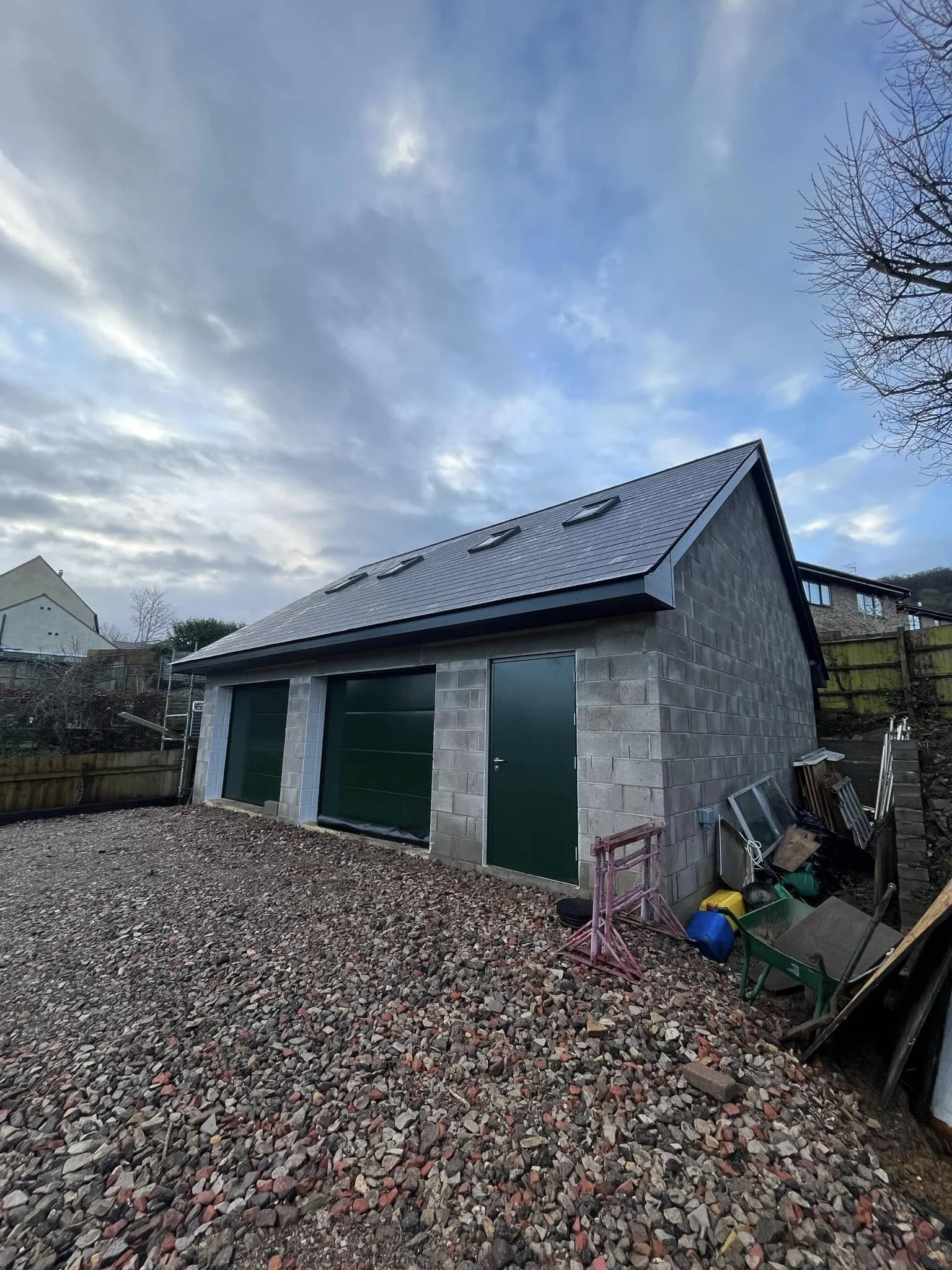 A small building with a sloped metal roof, two large green garage doors, a green side door, and small roof windows. The ground is covered in rocks, with various gardening tools and supplies nearby. The sky is partly cloudy with some sunlight.