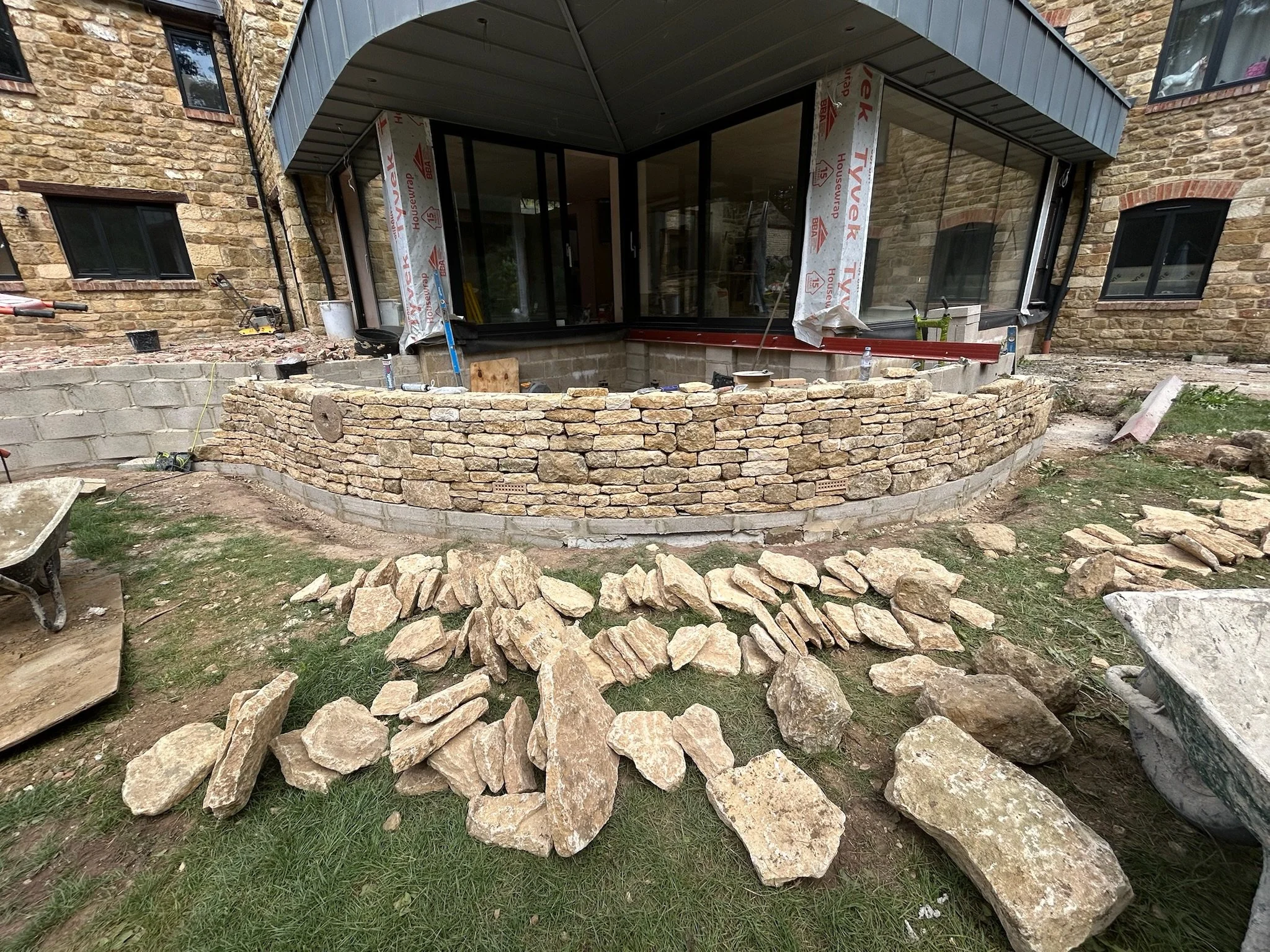 Construction site showing a building with a curved stone wall in progress, various construction tools and rocks scattered around, and a partly finished porch with scaffolding.